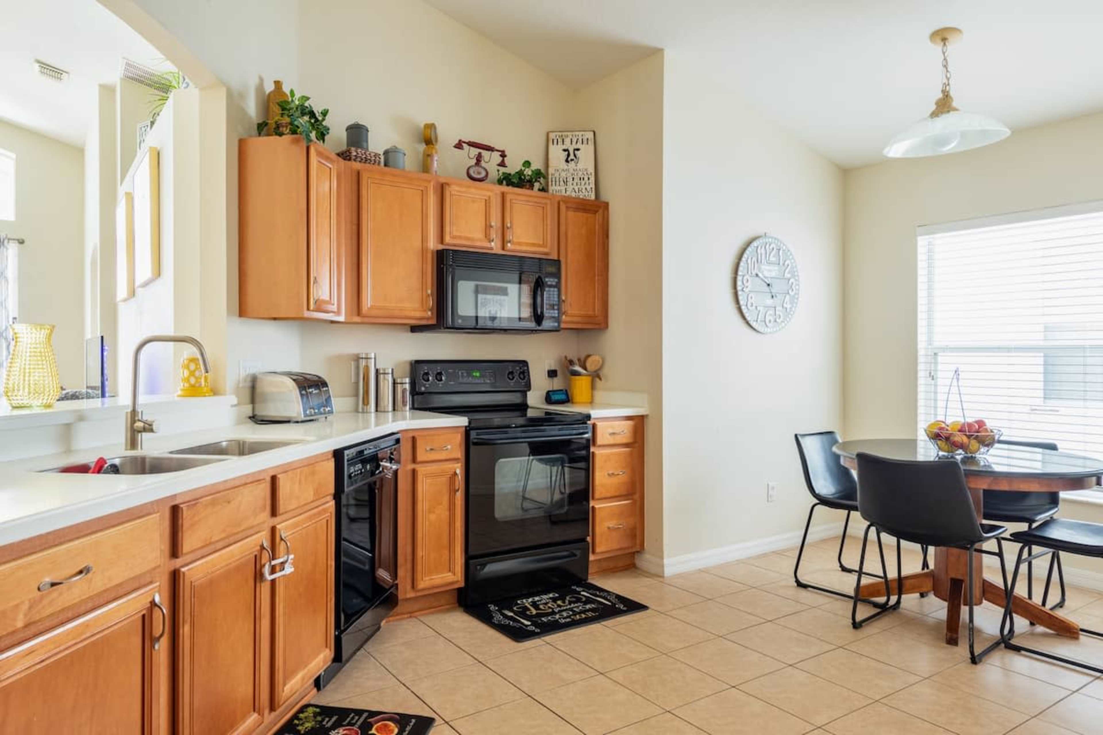 The image shows a modern kitchen with wooden cabinets, a black oven and microwave, and a dining area featuring a round table and chairs.