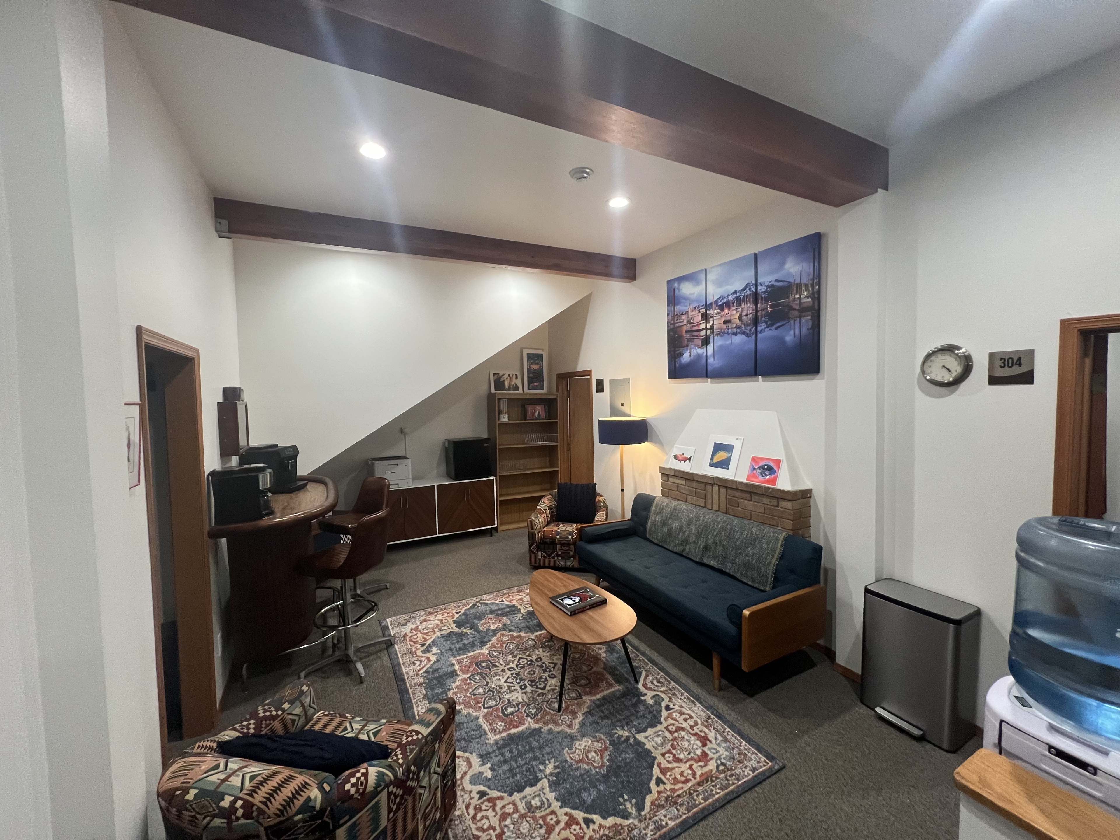 The image shows a cozy room with a sofa, armchair, coffee table, and a bar area along with various decorations, including wall art and a water cooler.