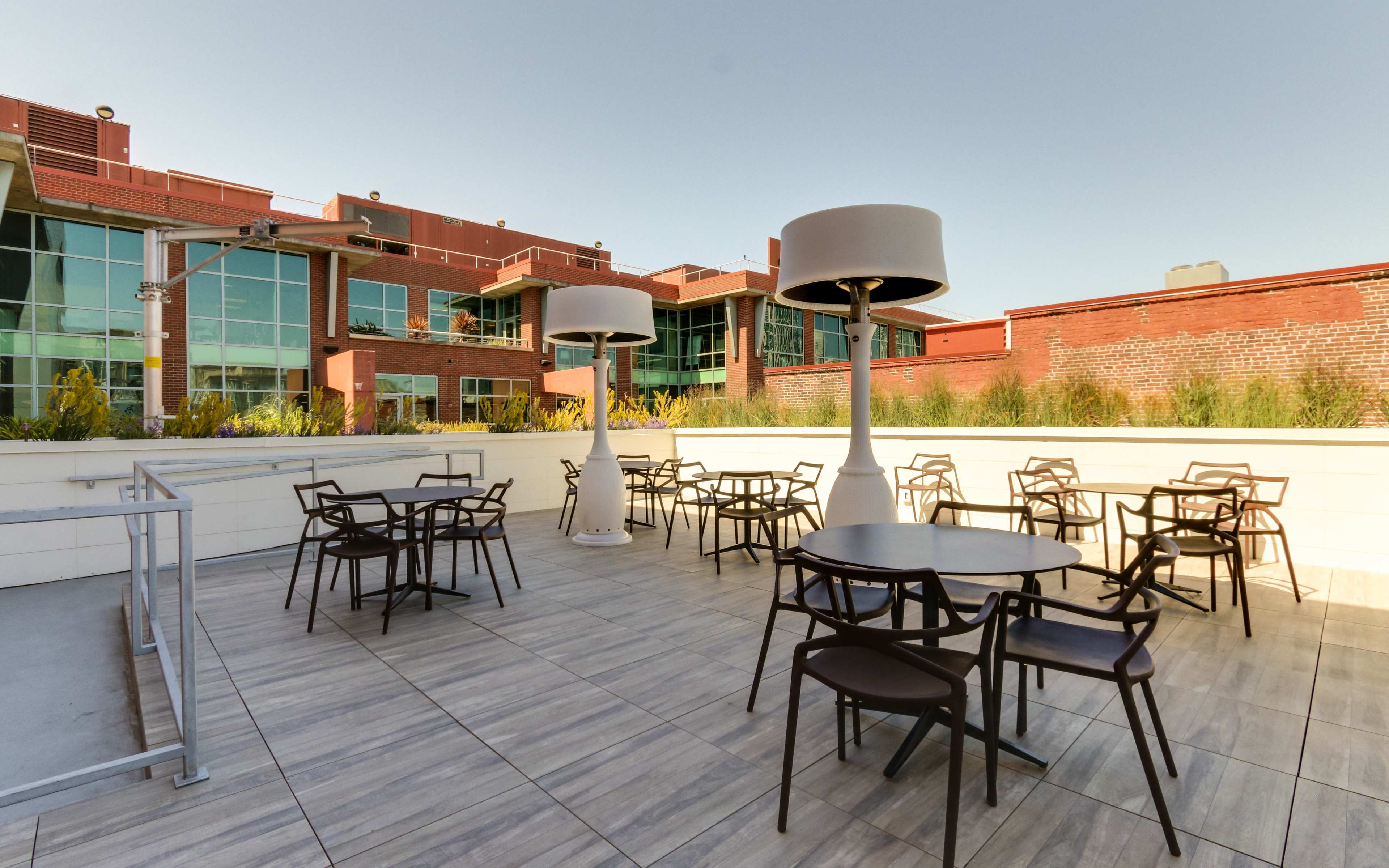 A rooftop terrace with several round tables and tall lamps surrounded by a modern building and greenery.