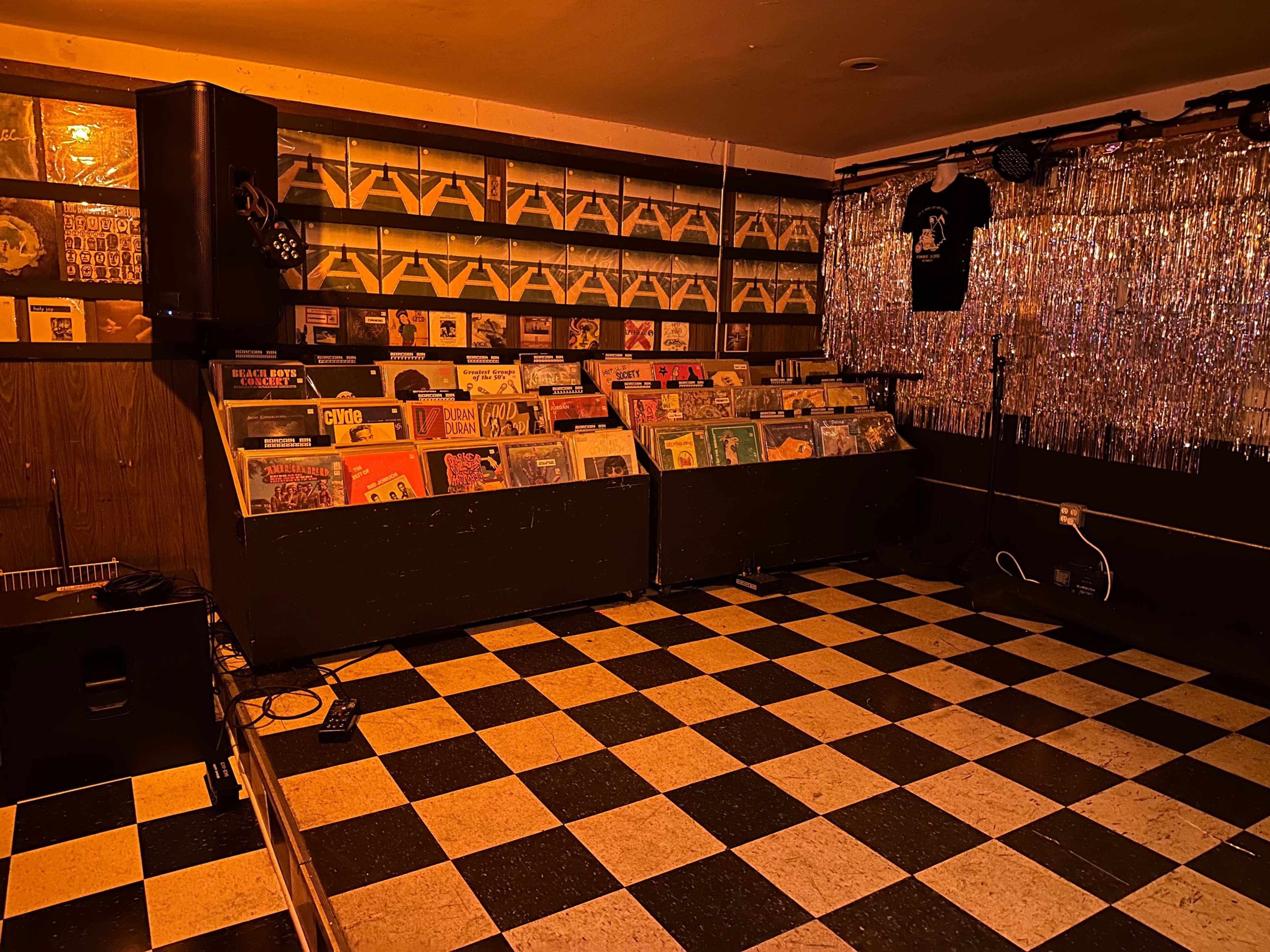 The image shows a small music venue with a checkered floor and a display of records in wooden crates along the walls.