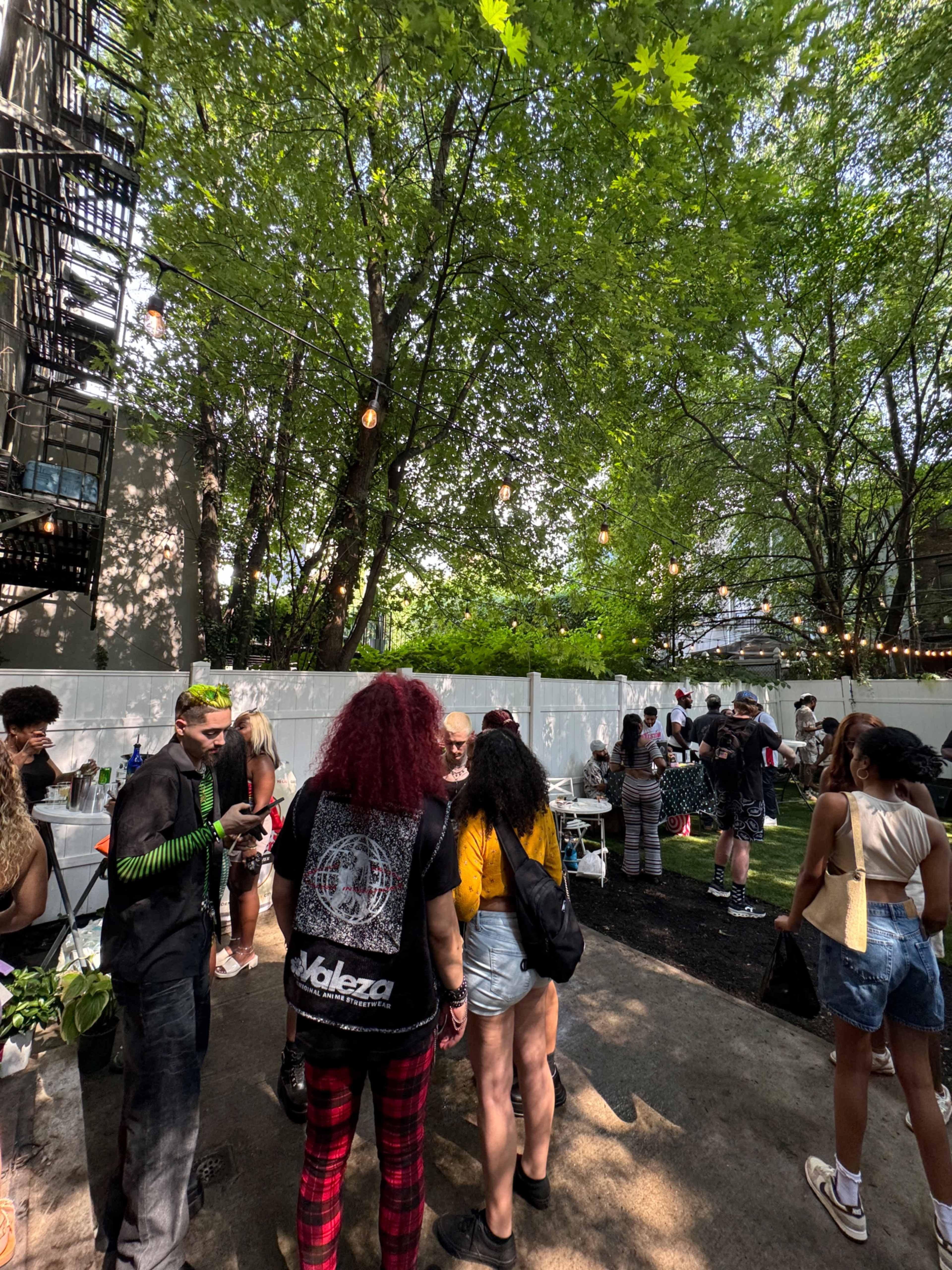A group of people socializes in a shaded outdoor area filled with trees and string lights.