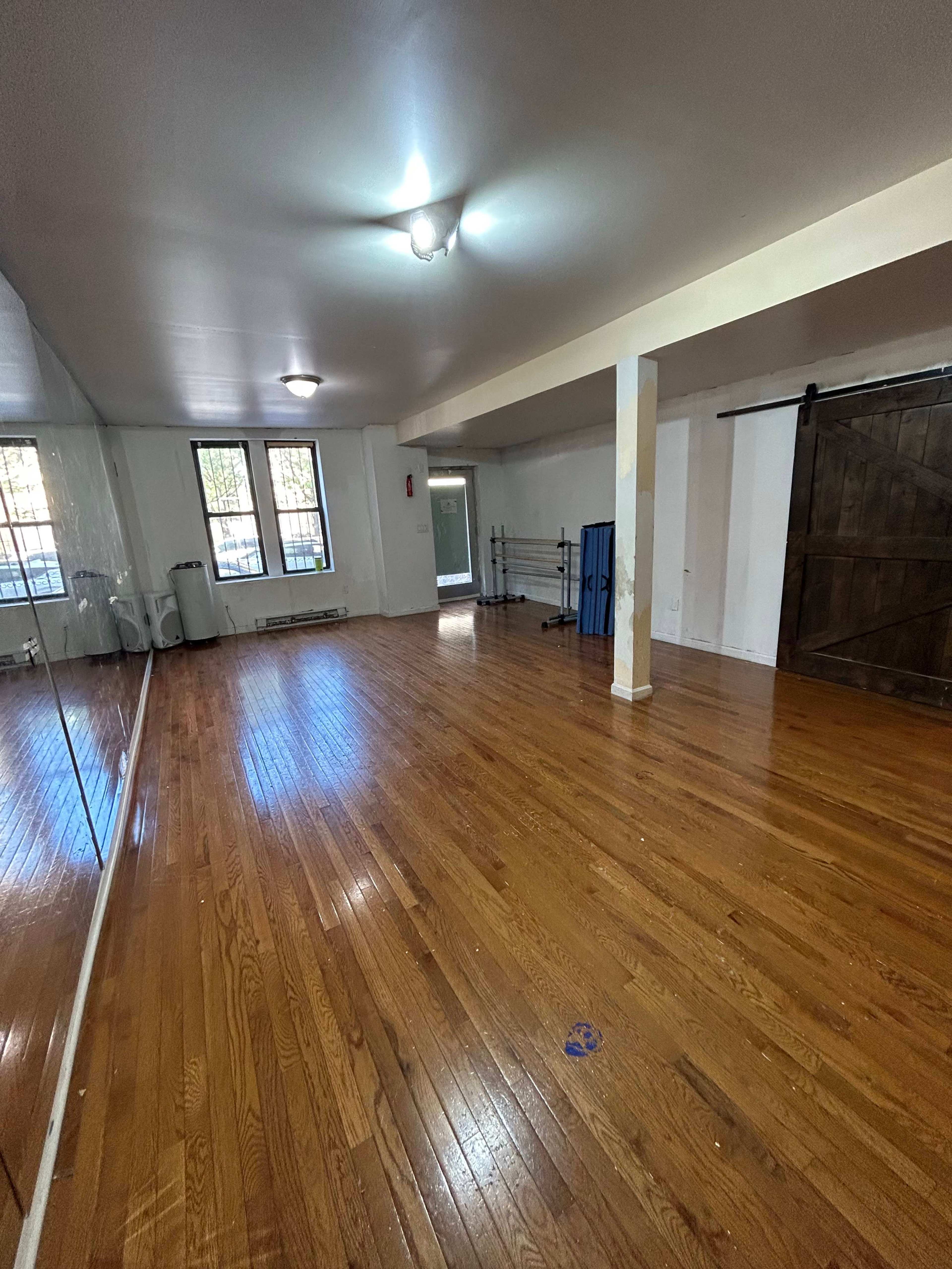 An empty room with hardwood flooring, large windows, and a mirrored wall on one side.