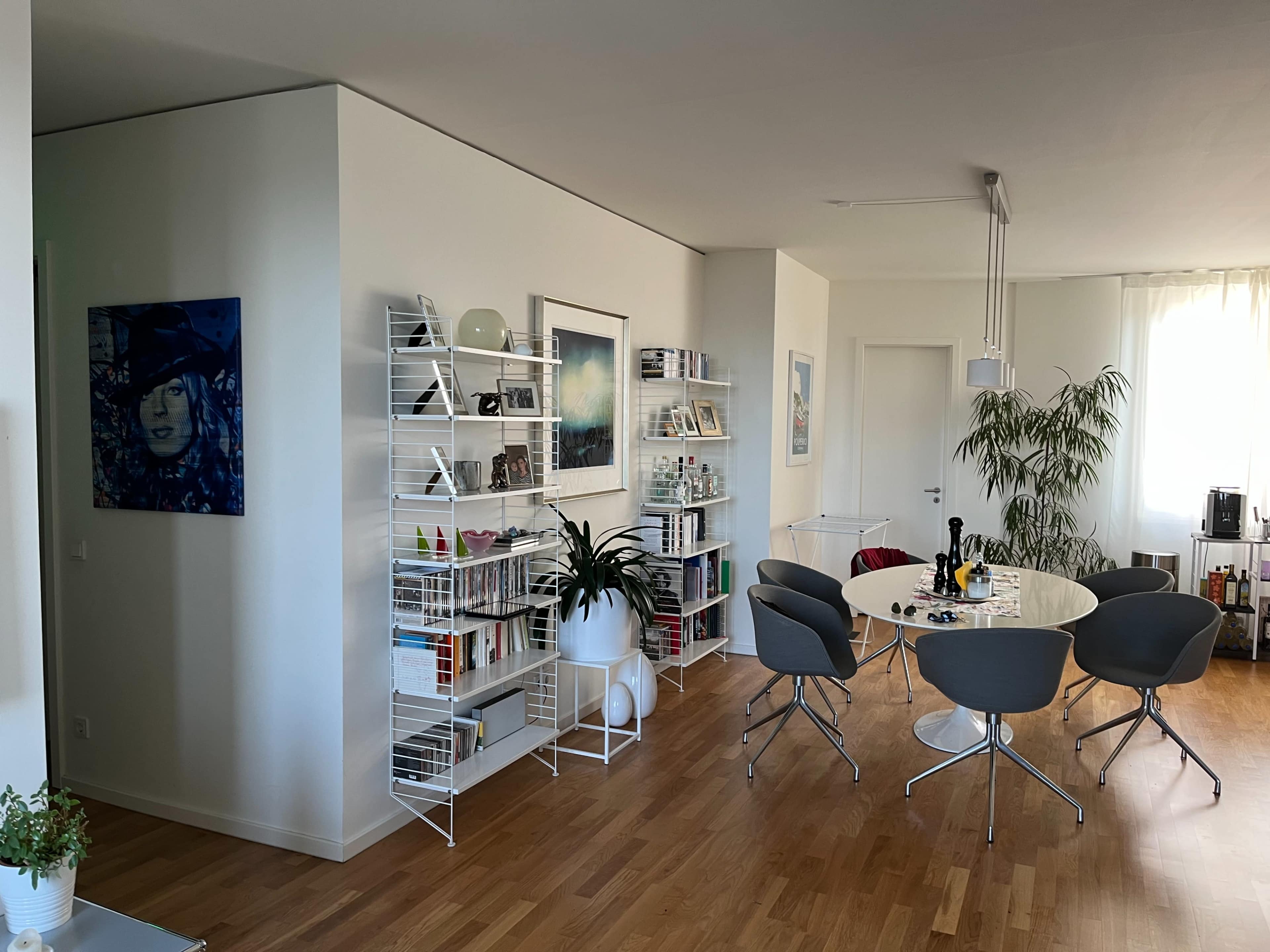 The image shows a modern living space with a round dining table surrounded by four chairs, a bookshelf filled with books, and artwork on the walls.