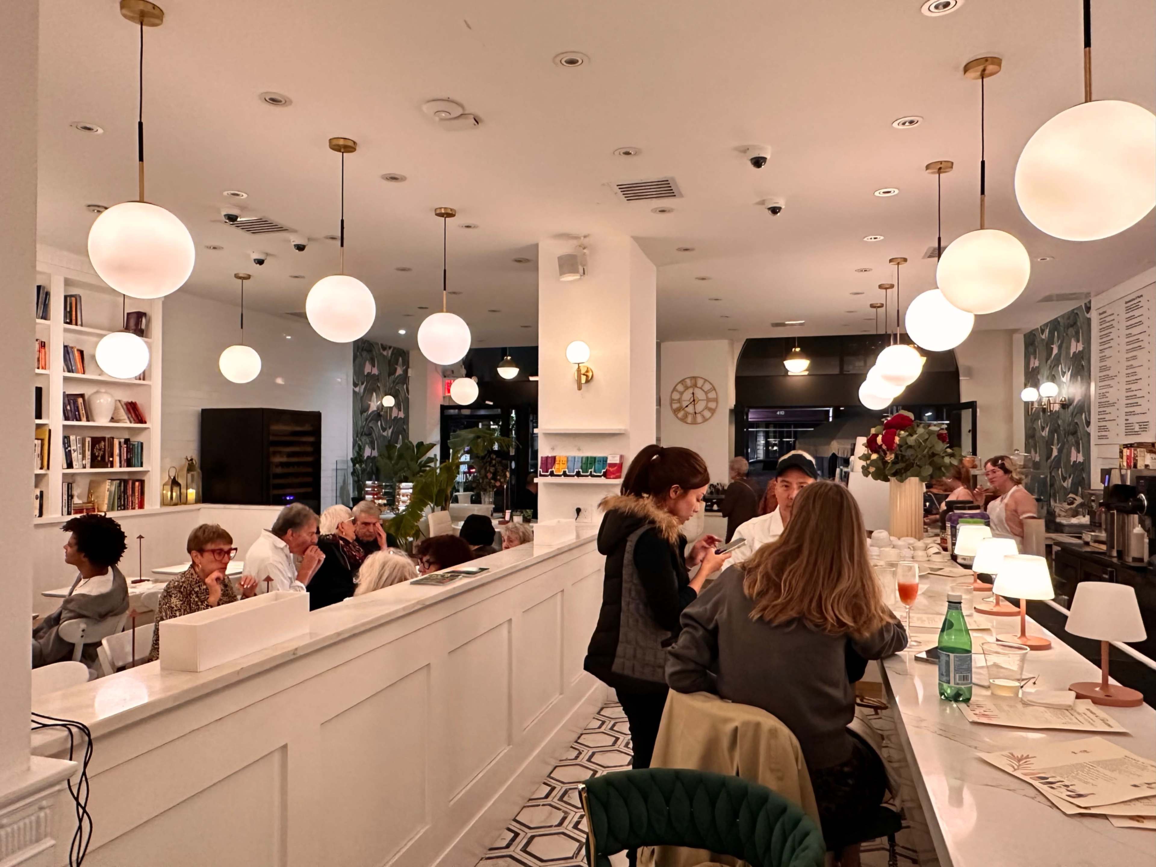 The scene features a modern café filled with patrons seated at tables and counters, illuminated by round pendant lights and decorated with plants and bookshelves.