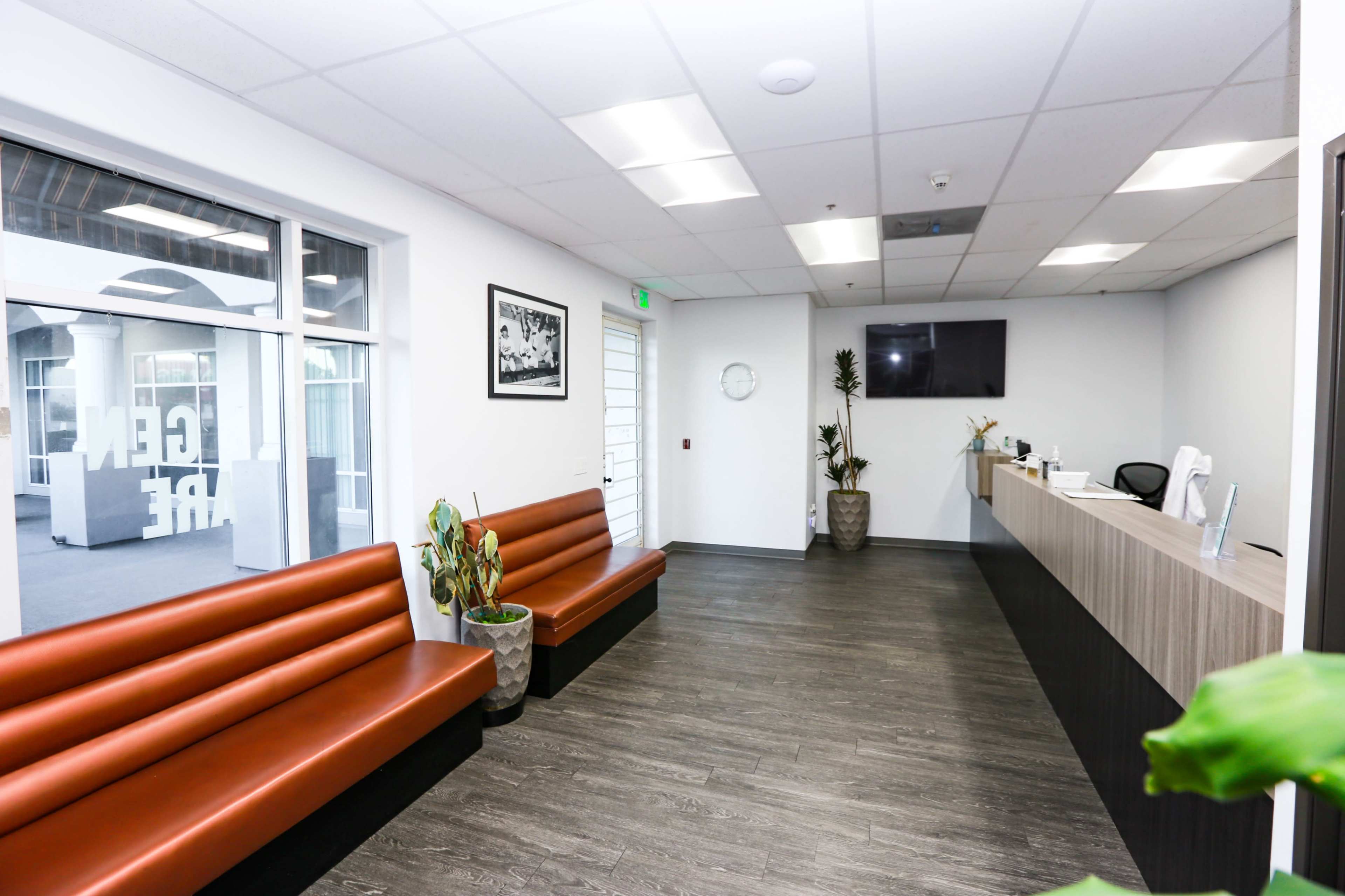 The image shows a modern reception area with two brown benches, a front desk, and decorative plants in pots.