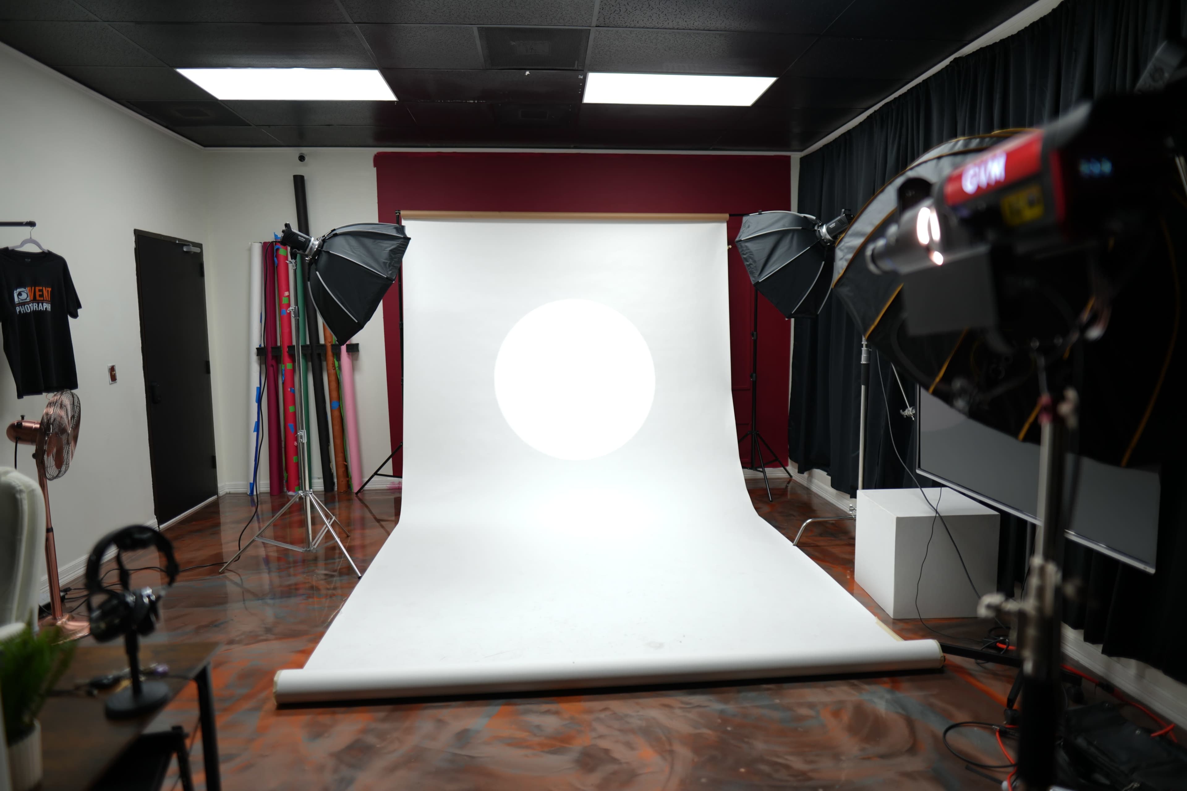 The image shows a photography studio with a white backdrop, two softbox lights on either side, and various photography equipment arranged around the space.