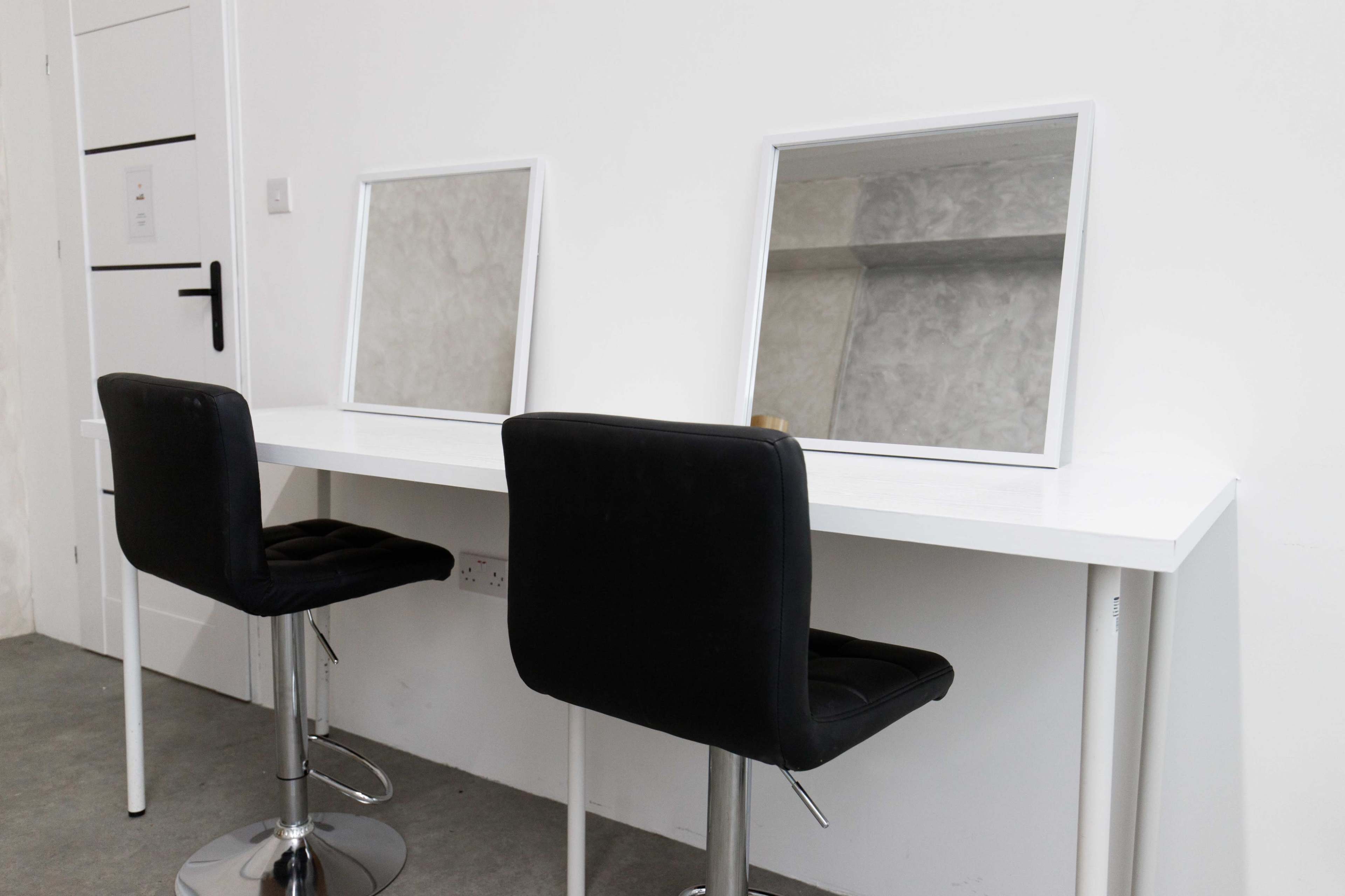 The image shows a white table with two square mirrors facing each other, accompanied by two black, padded stools.