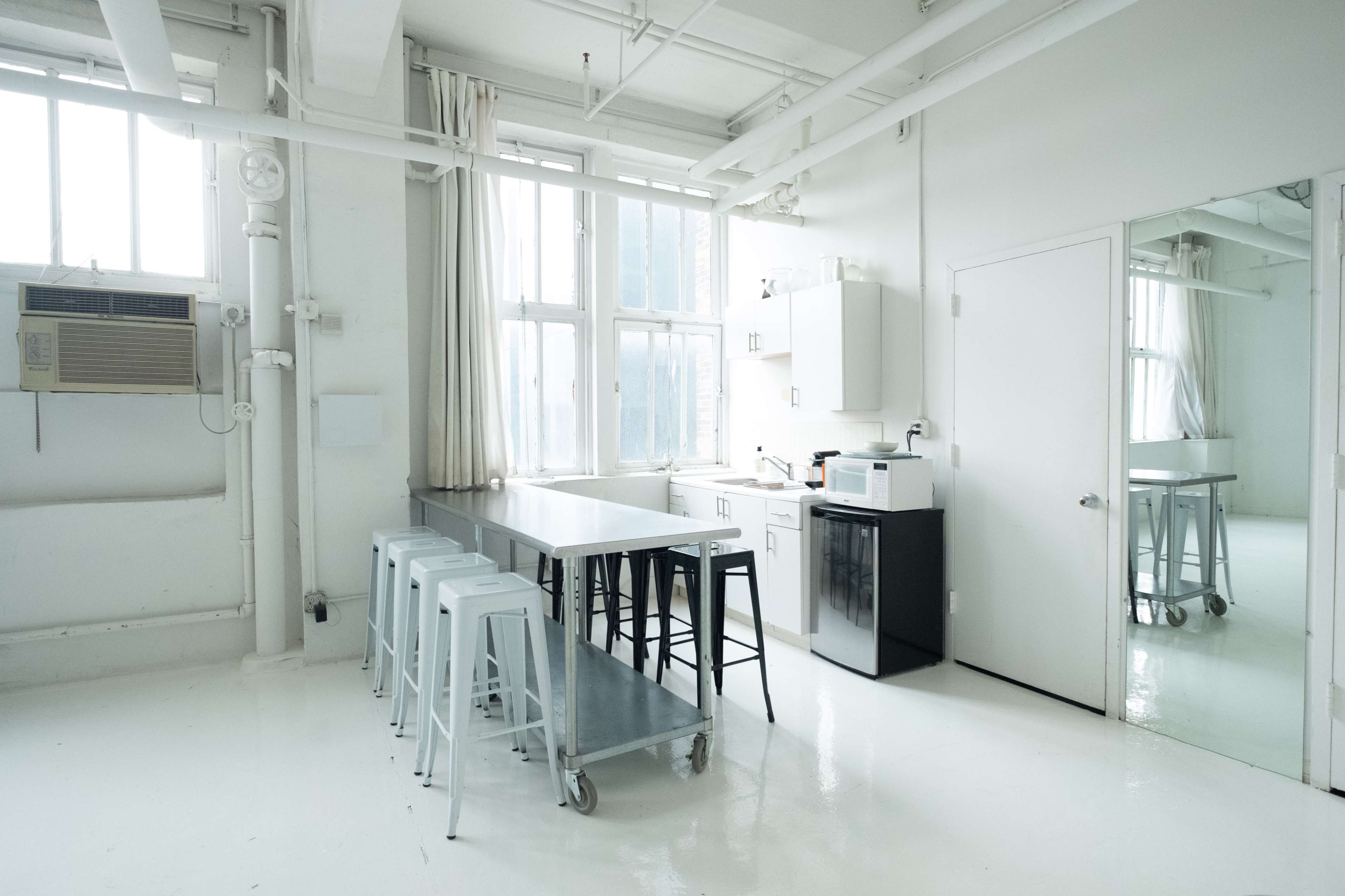The image shows a modern kitchen with a long table, metal stools, a refrigerator, and large windows allowing natural light to enter.