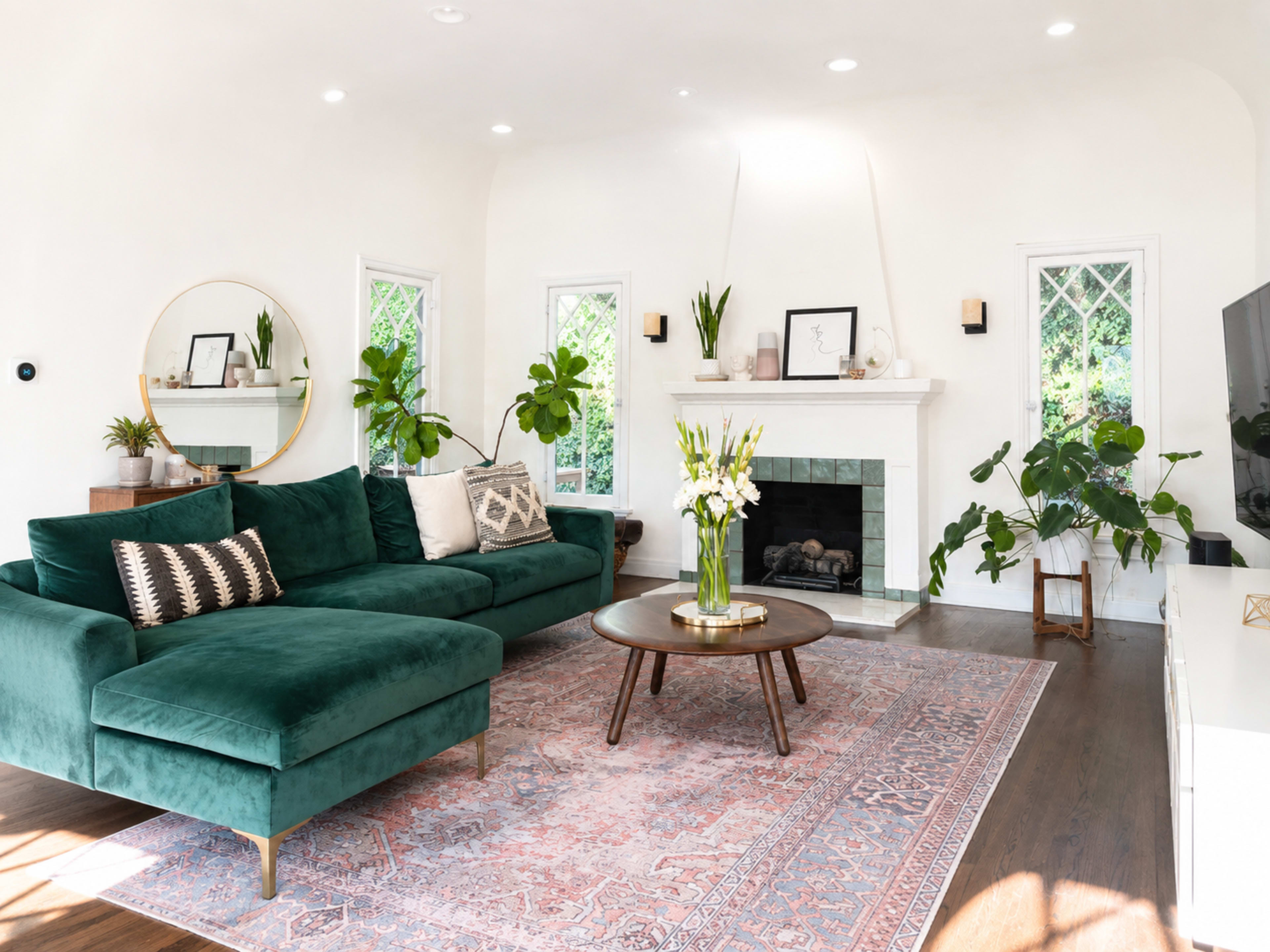 The living room features a green sectional sofa, a round coffee table, and potted plants, with a fireplace and a large mirror enhancing the space.