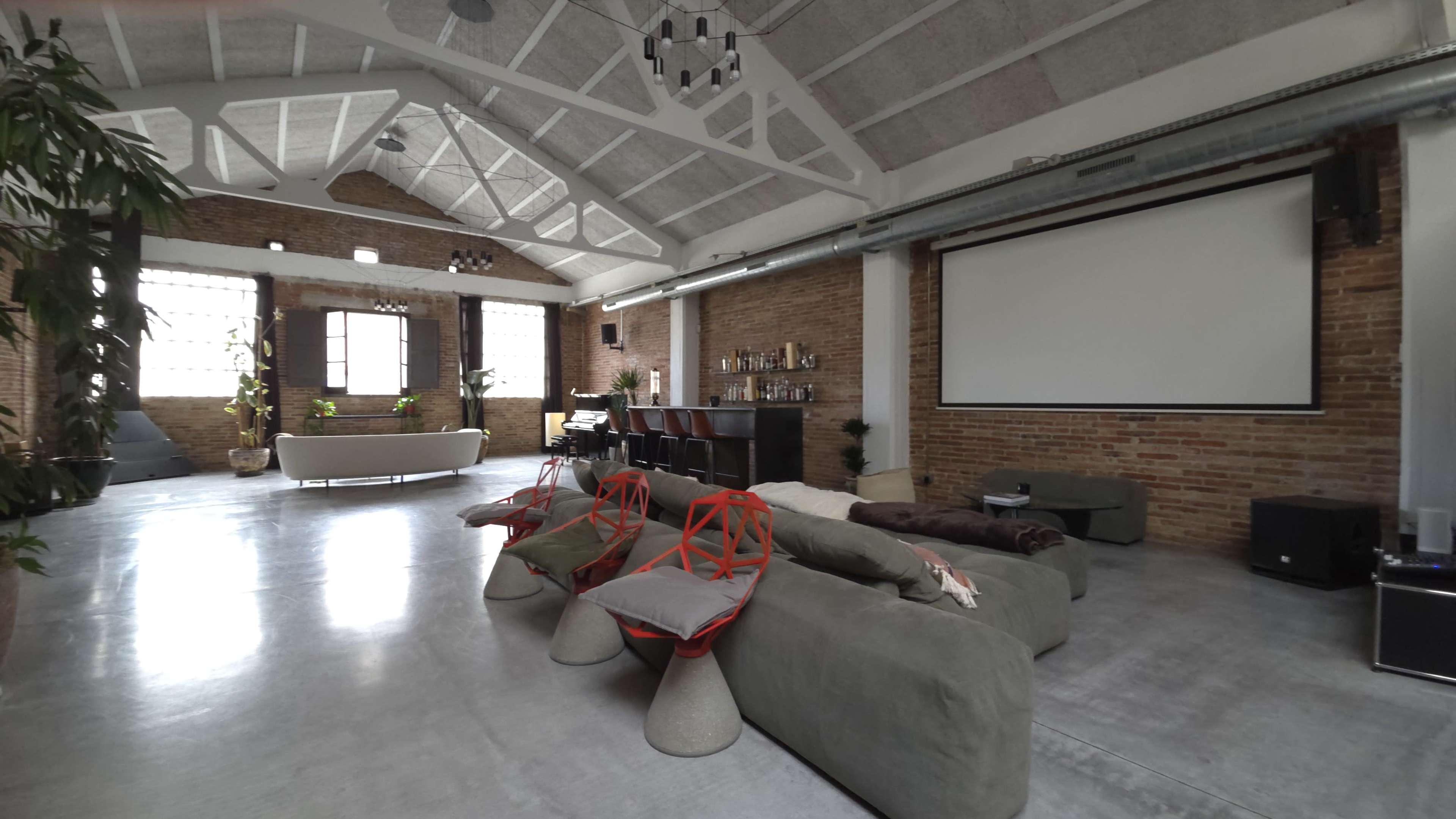 The image shows a spacious industrial-style room with exposed brick walls, large windows, and a projection screen, featuring modern seating arrangements and a bar area.