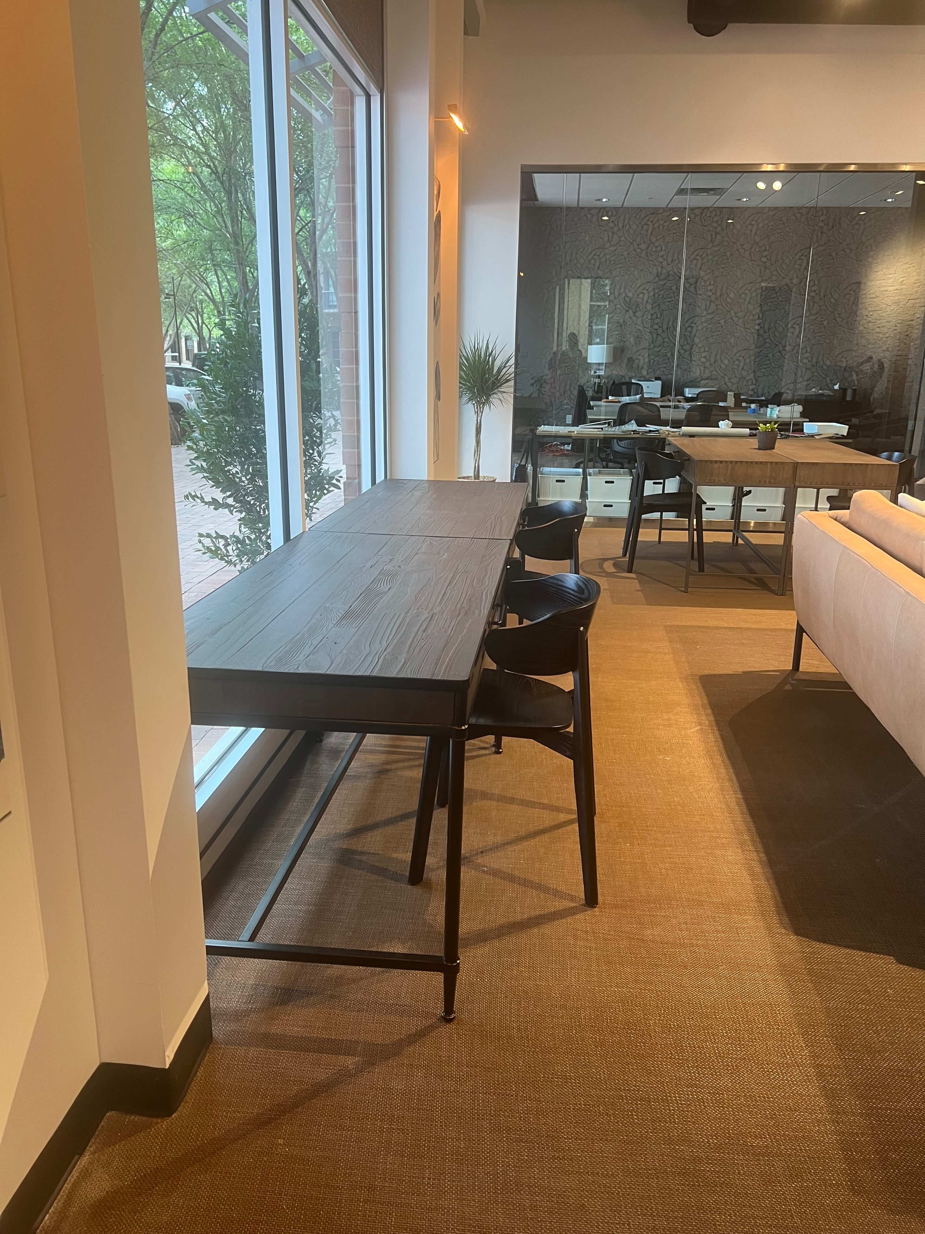 A long wooden table with black chairs is positioned next to a large window in a modern office space.