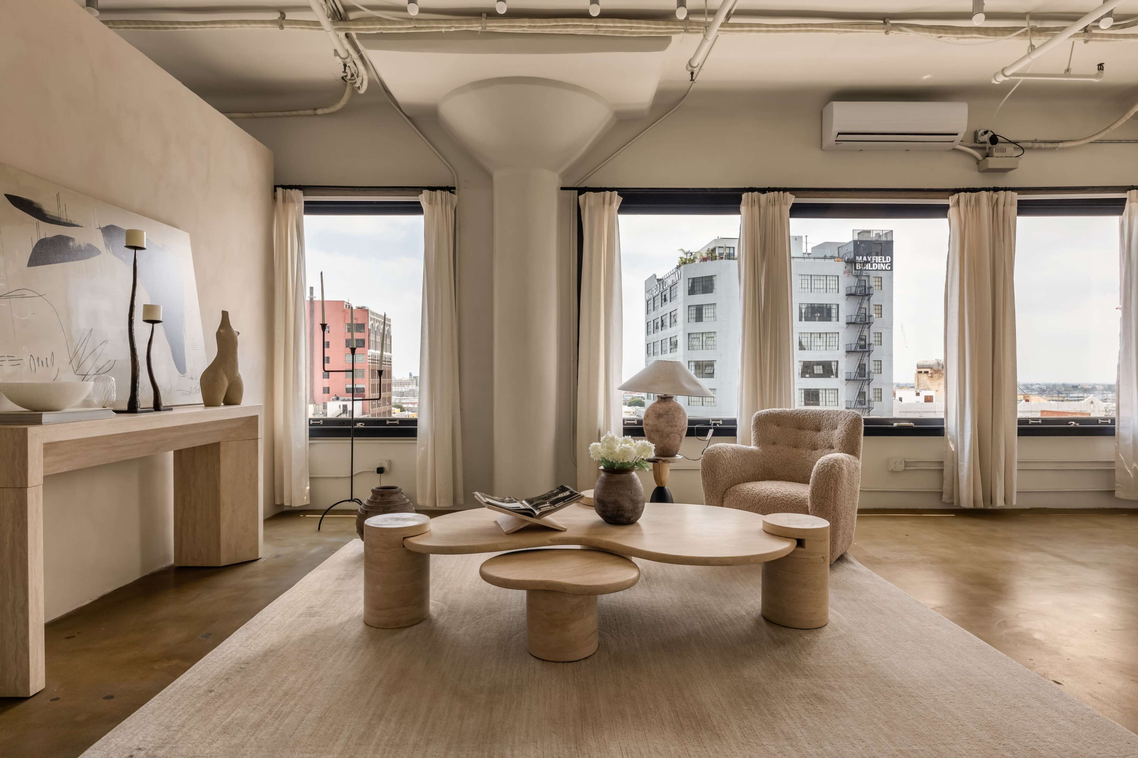 The image shows a modern living room with a round wooden coffee table, a beige armchair, and large windows showcasing a city view.