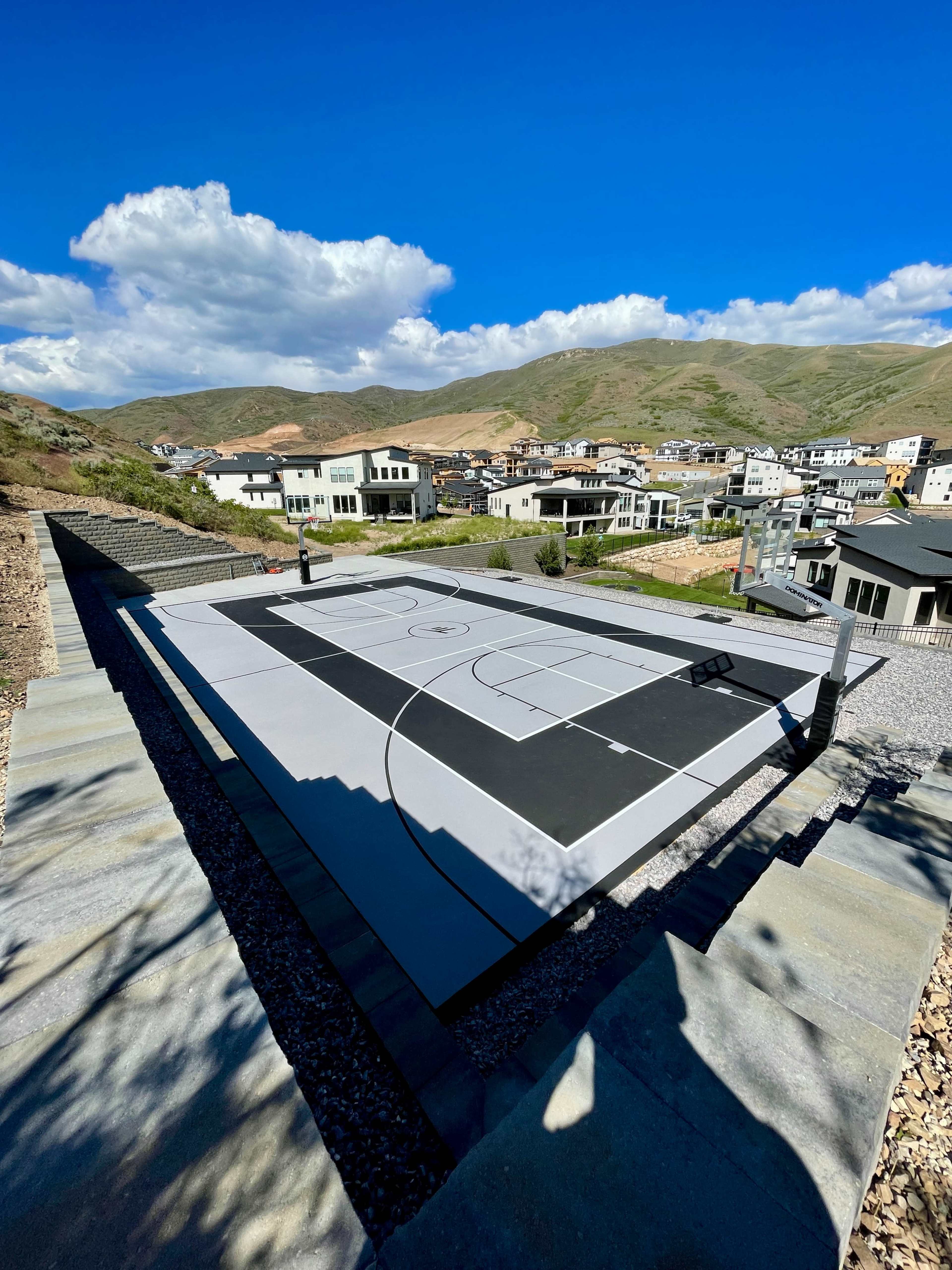A basketball court is situated on a hilltop, surrounded by modern houses and a mountainous landscape under a clear blue sky.