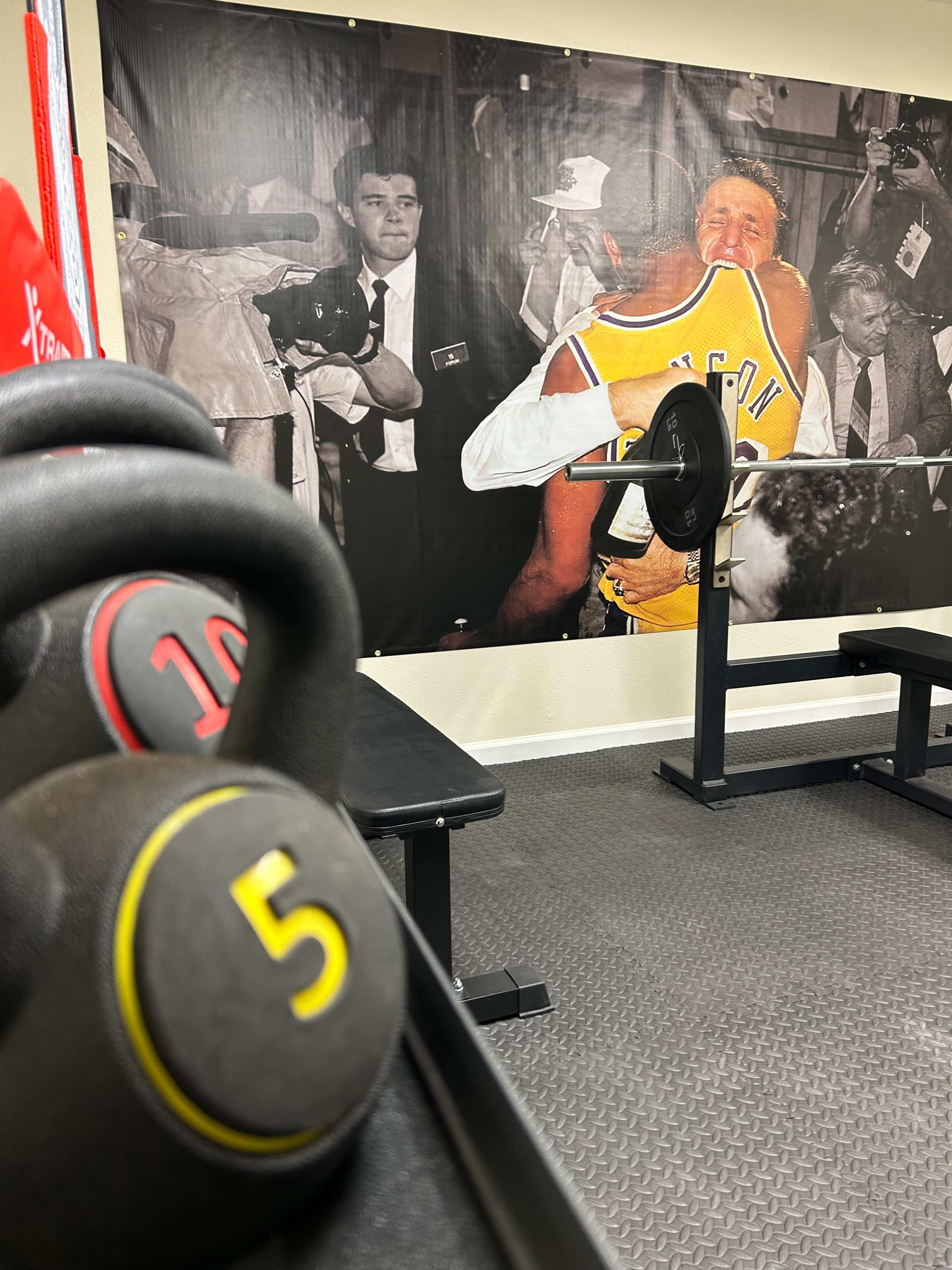 The image shows a gym interior with a focus on a set of dumbbells in the foreground and a large mural of two athletes embracing in the background.