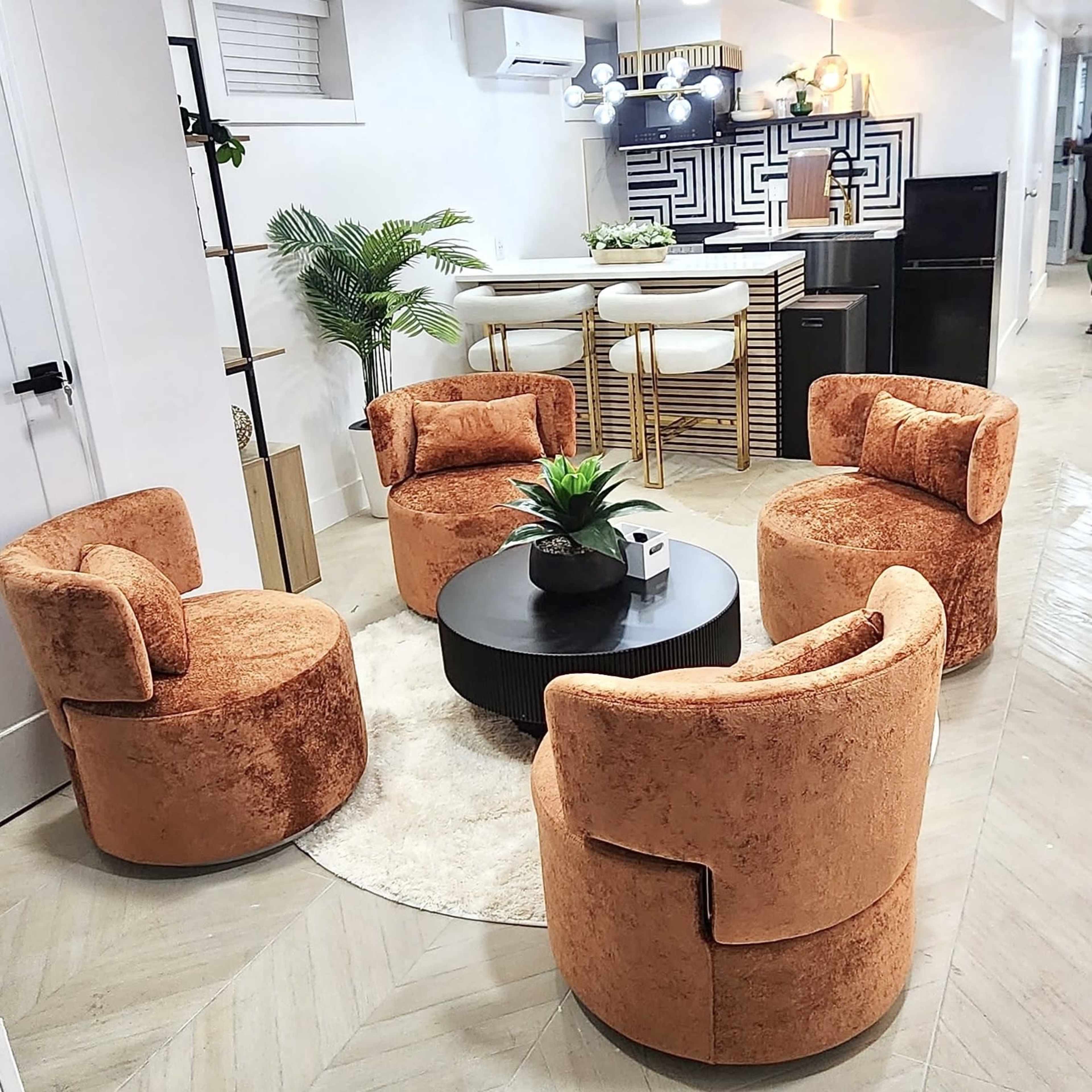 The image shows a modern living space featuring four round, orange upholstered chairs arranged around a circular black coffee table on a light rug.