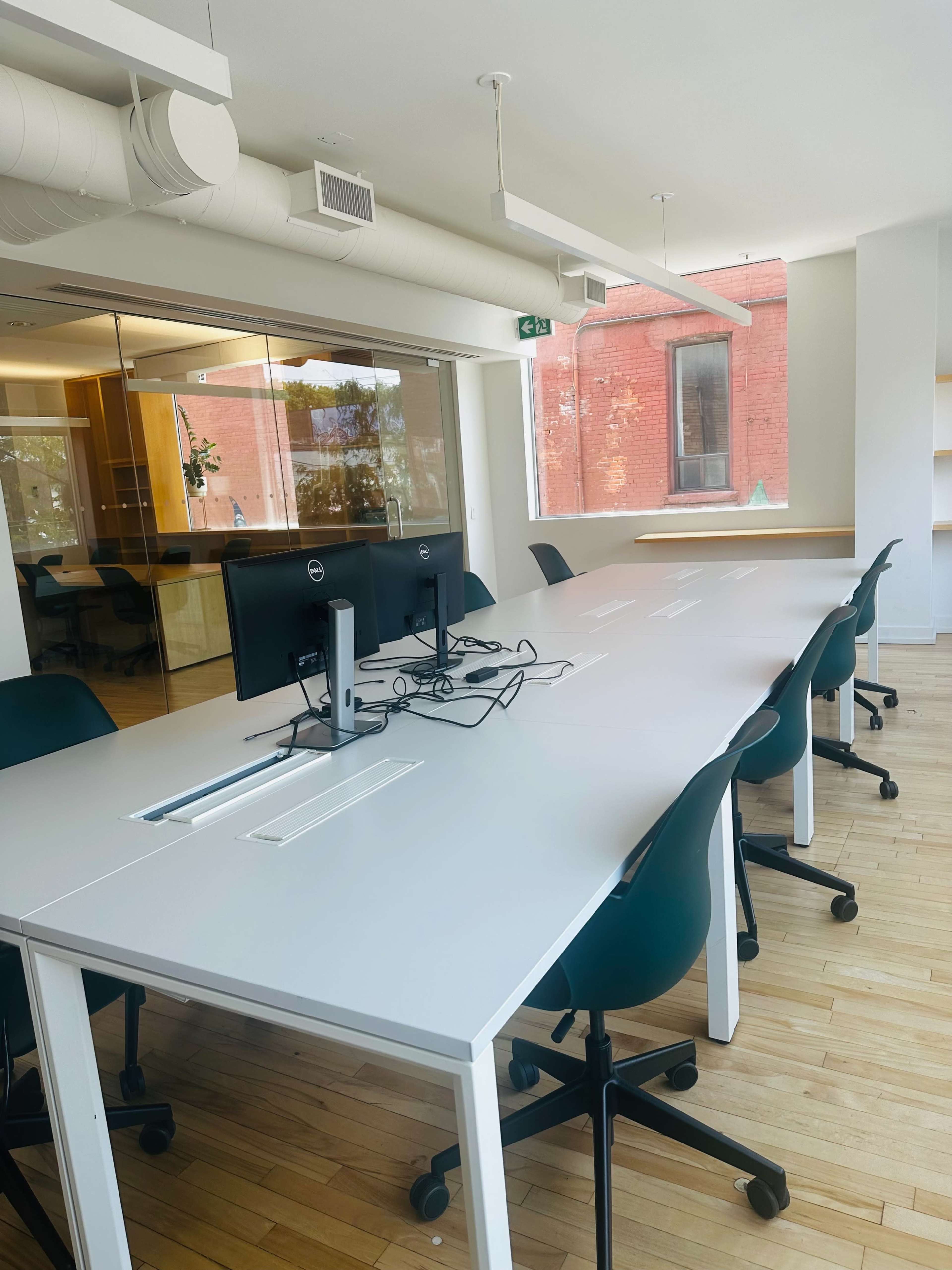 The image shows a modern office space featuring a long white conference table with two computer monitors and green chairs, illuminated by natural light from large windows.