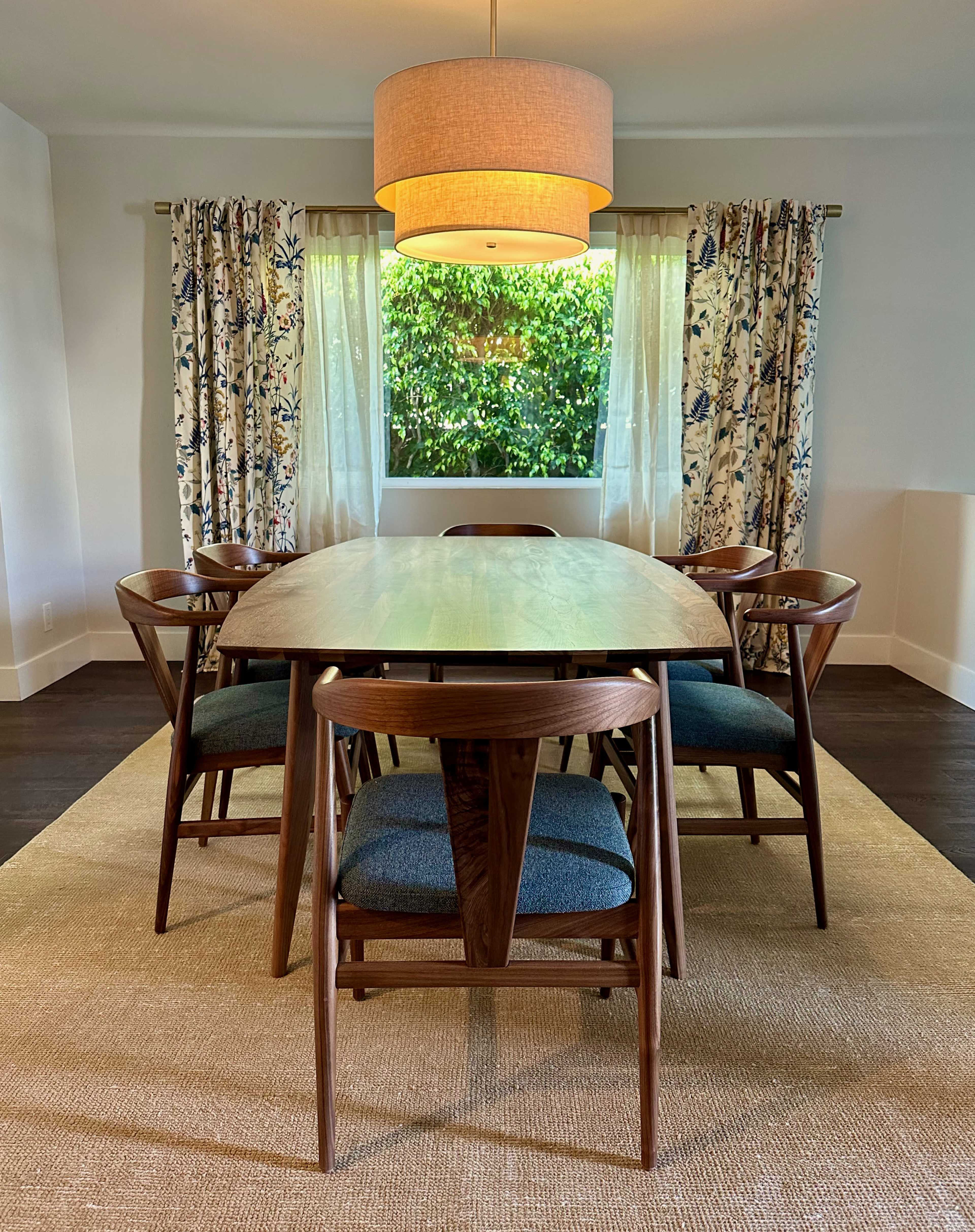 A wooden dining table surrounded by six chairs is illuminated by a hanging light fixture, with drapes framing a window that overlooks greenery.