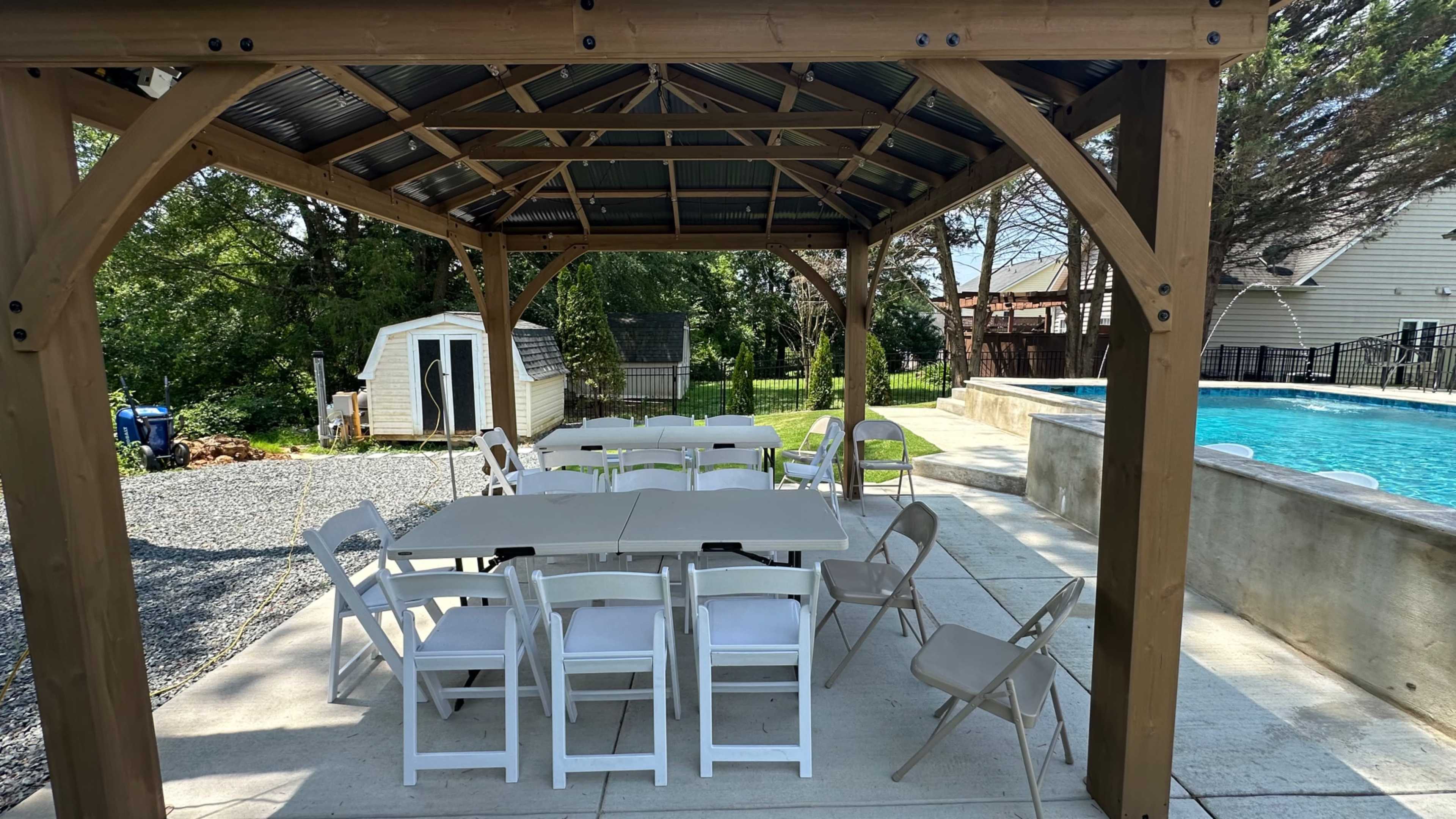 A covered outdoor pavilion filled with white tables and chairs is situated beside a swimming pool and a well-maintained lawn.