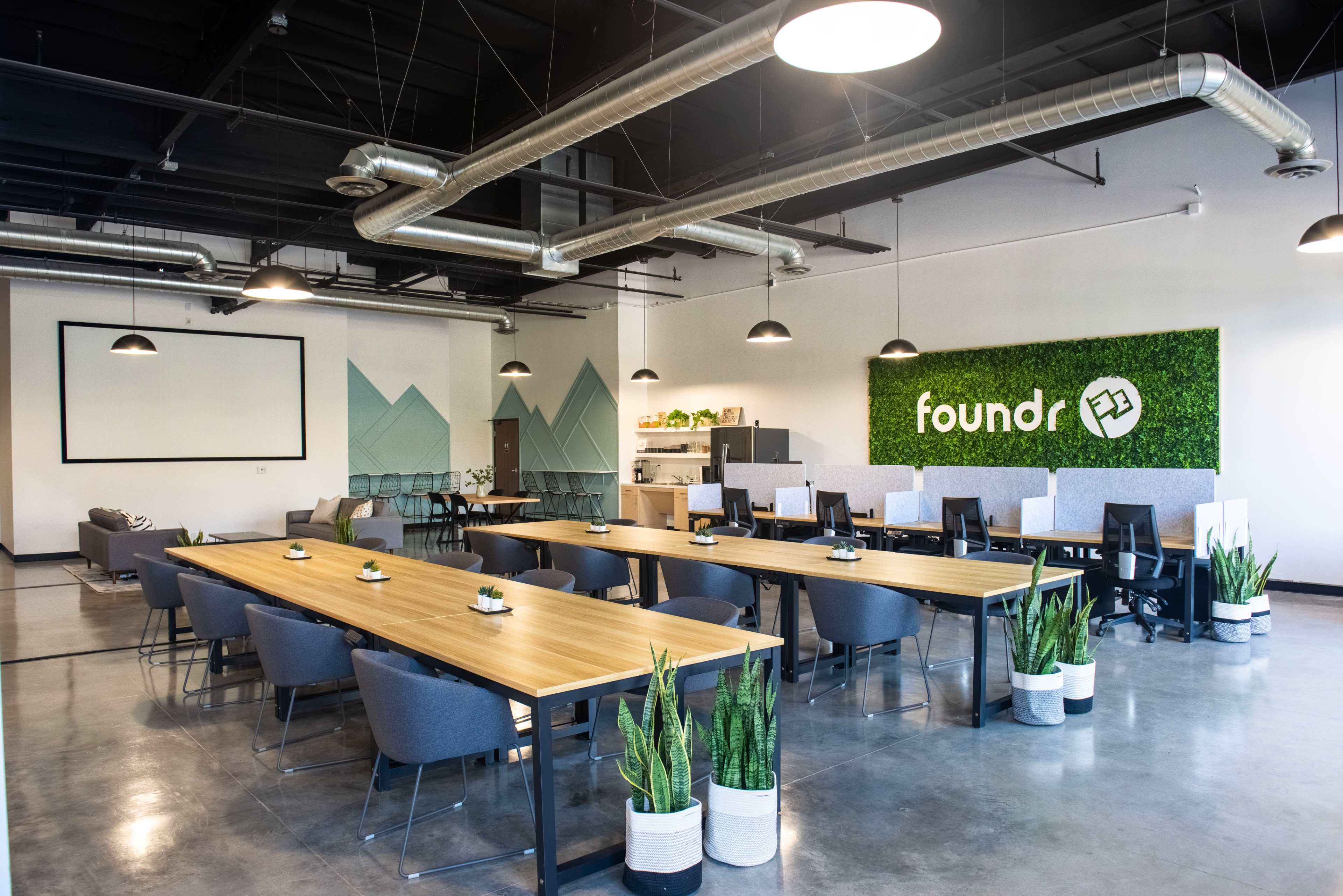 A modern office space with multiple workstations, large tables, and greenery decor on the walls.