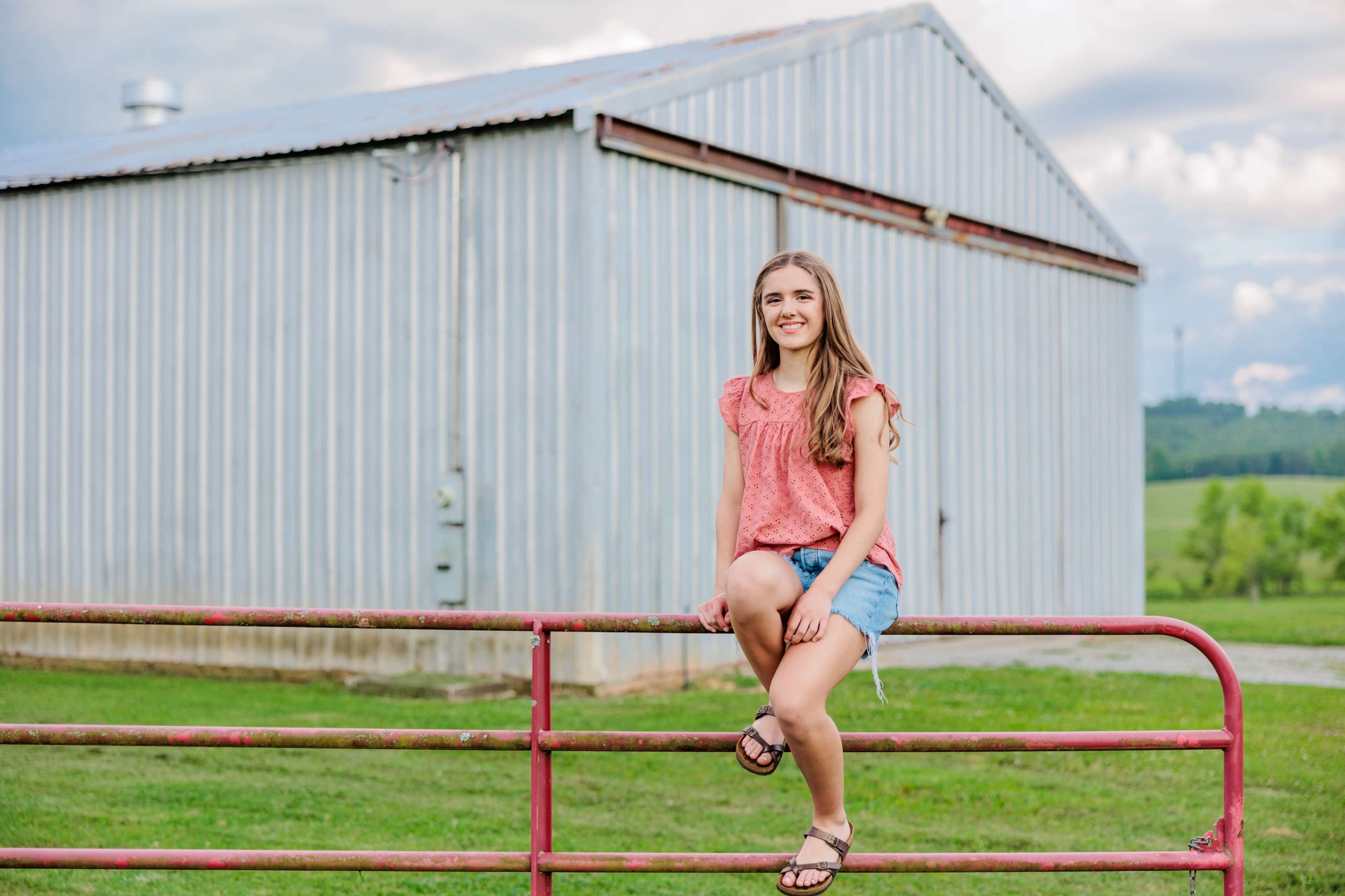 A girl sits on a pink fence in front of a metallic barn, with green fields and a cloudy sky visible in the background.