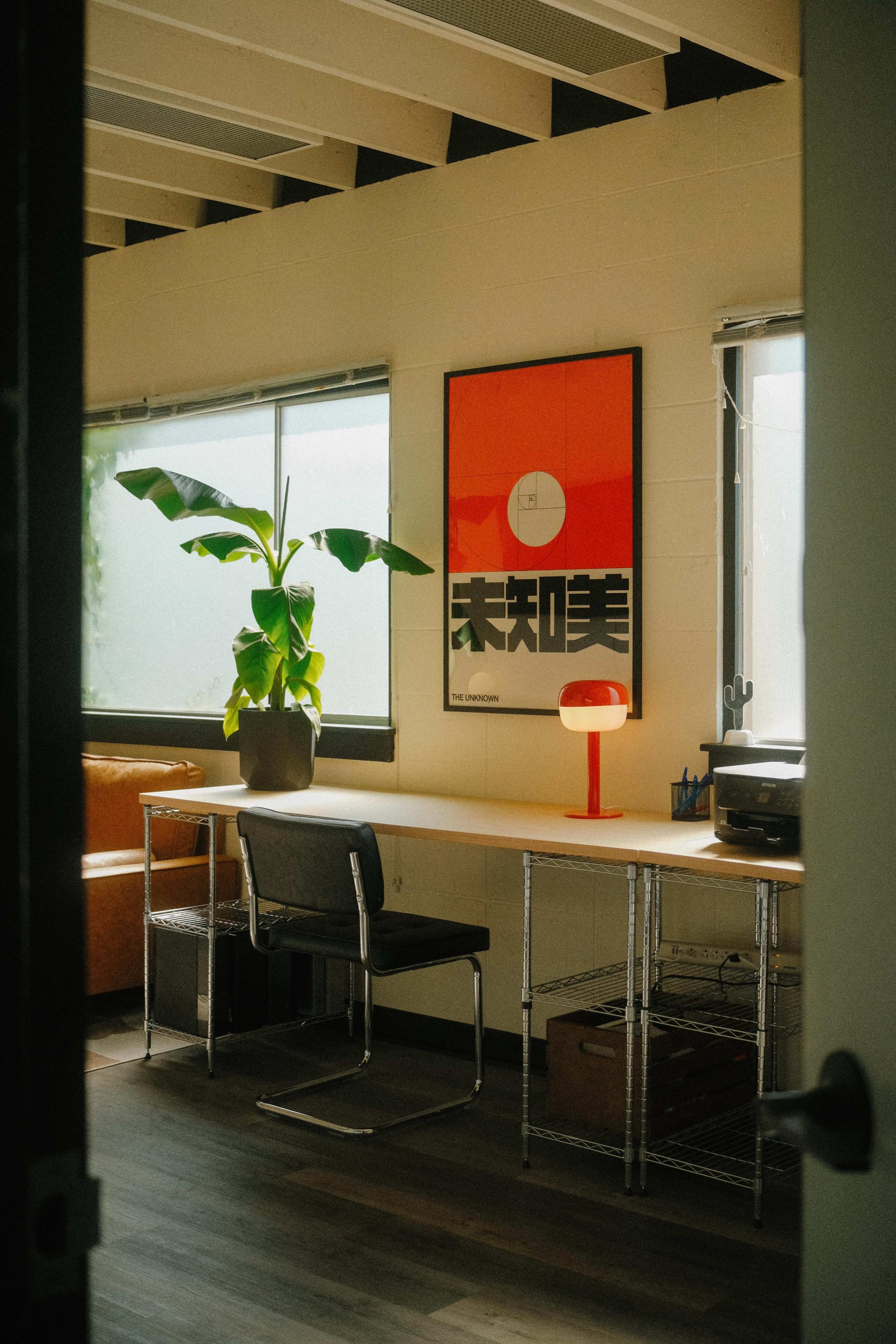 A modern office space features a desk with a potted plant, a chair, and a red lamp, accented by a large poster on the wall and large windows providing natural light.