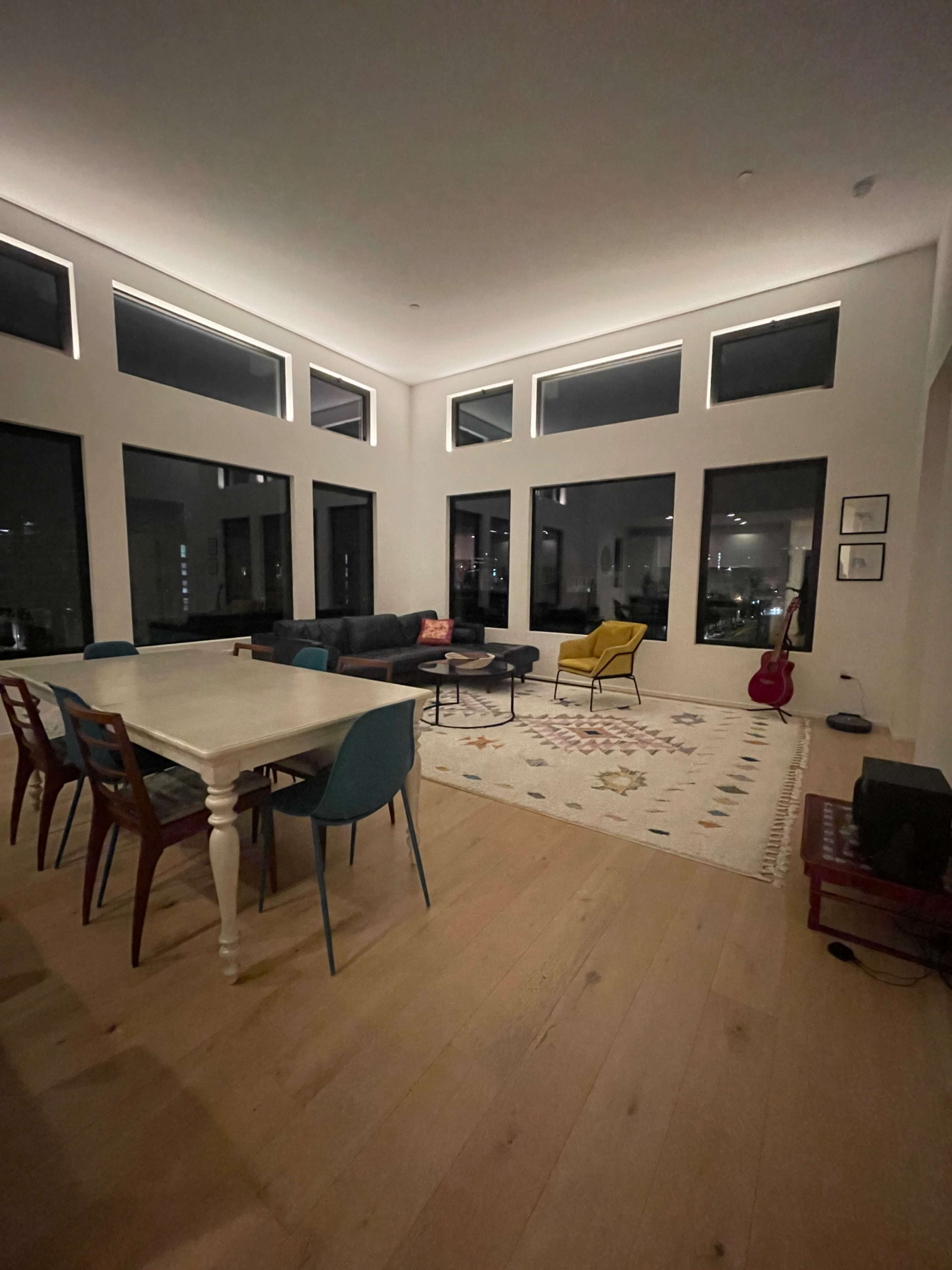 A modern living space features a dining table with colorful chairs, a dark sectional couch, a yellow armchair, and large windows allowing light to enter.