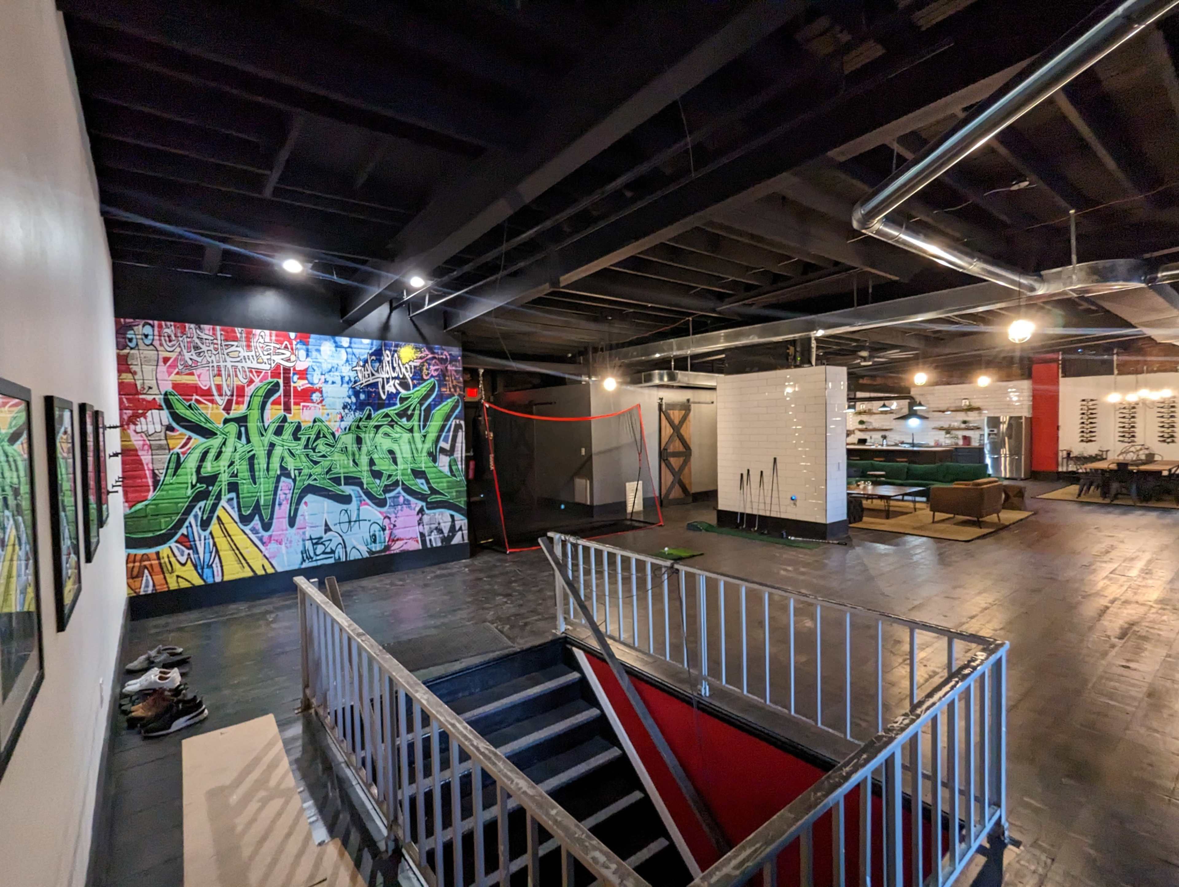 A spacious interior with a staircase leading down, a colorful graffiti wall, and various sections including a kitchen area and pool tables.