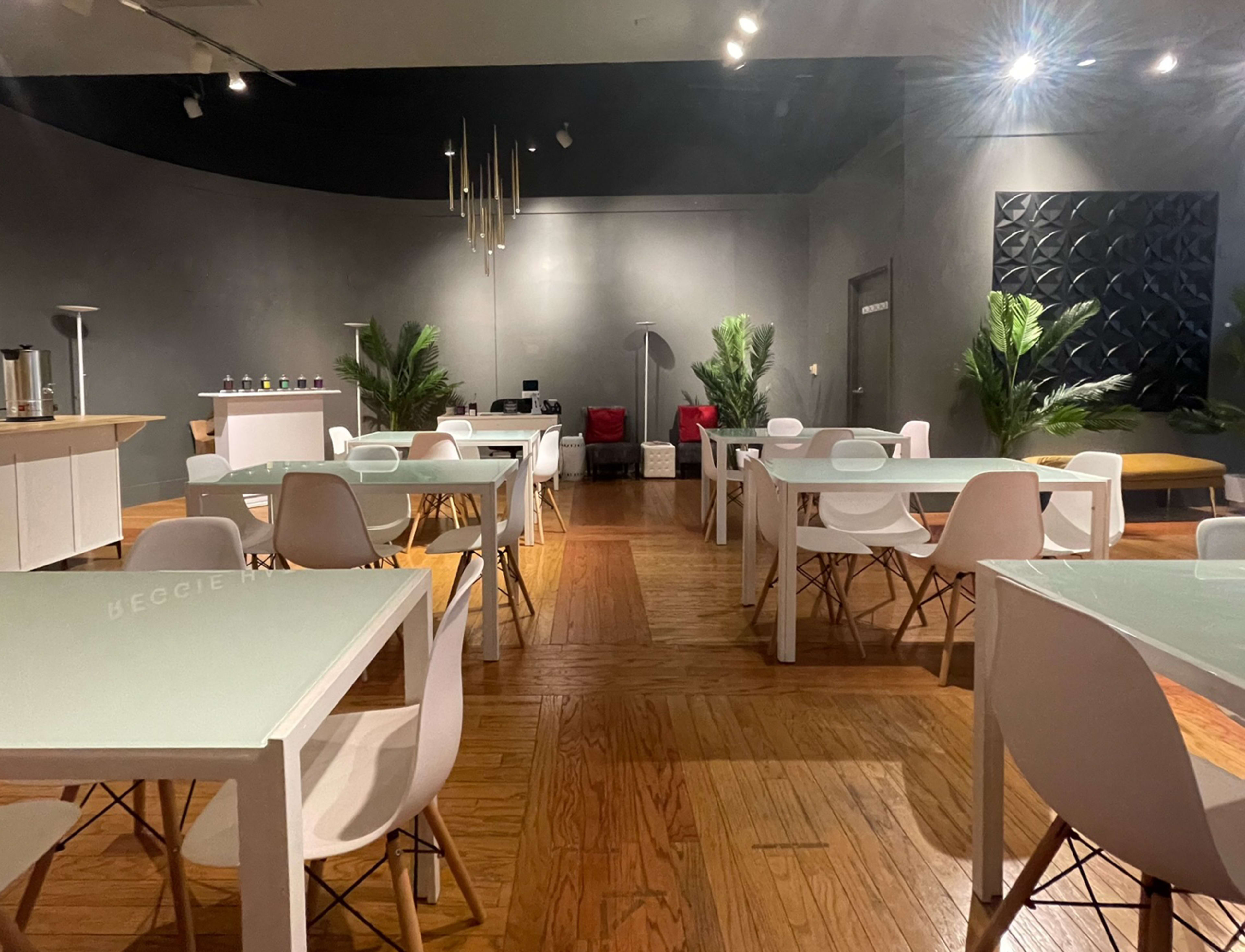 A modern, sparsely furnished interior with white tables and chairs, wooden flooring, and a few decorative plants.