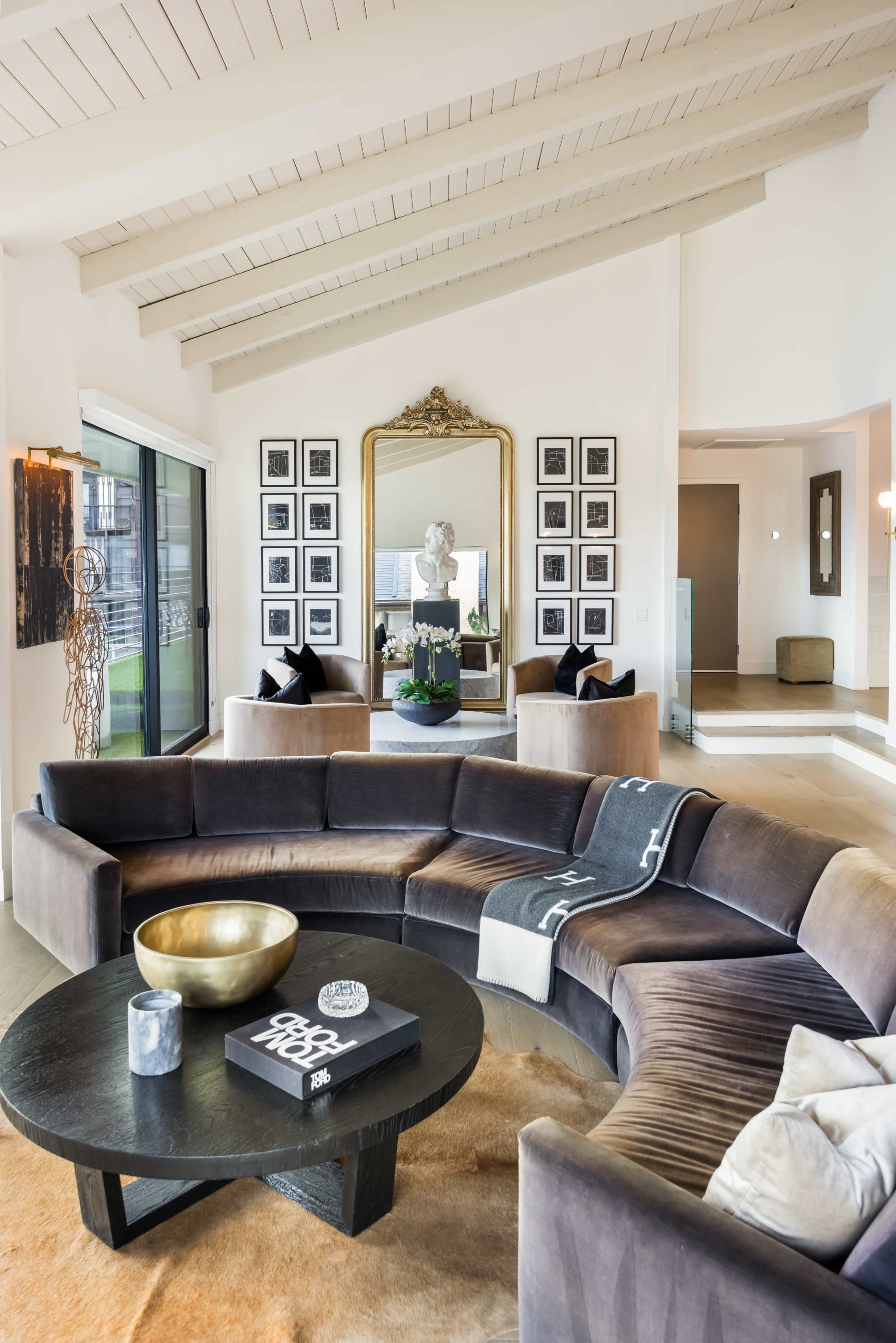 The image depicts a modern living room featuring a curved brown sofa, a round black coffee table, and a large mirror surrounded by a grid of framed black-and-white photos.