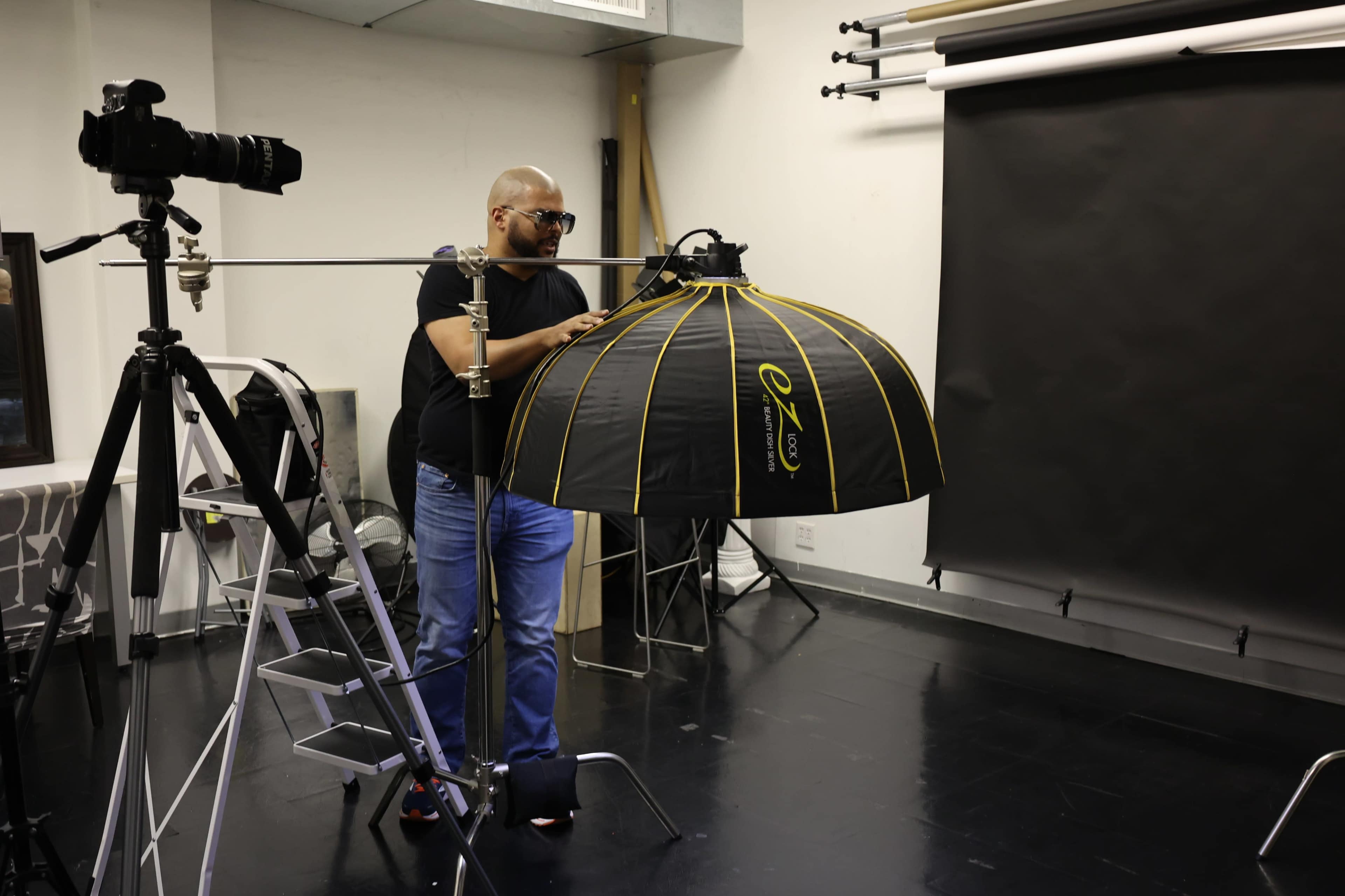 A man adjusts a large softbox light in a photography studio equipped with a camera and a backdrop.