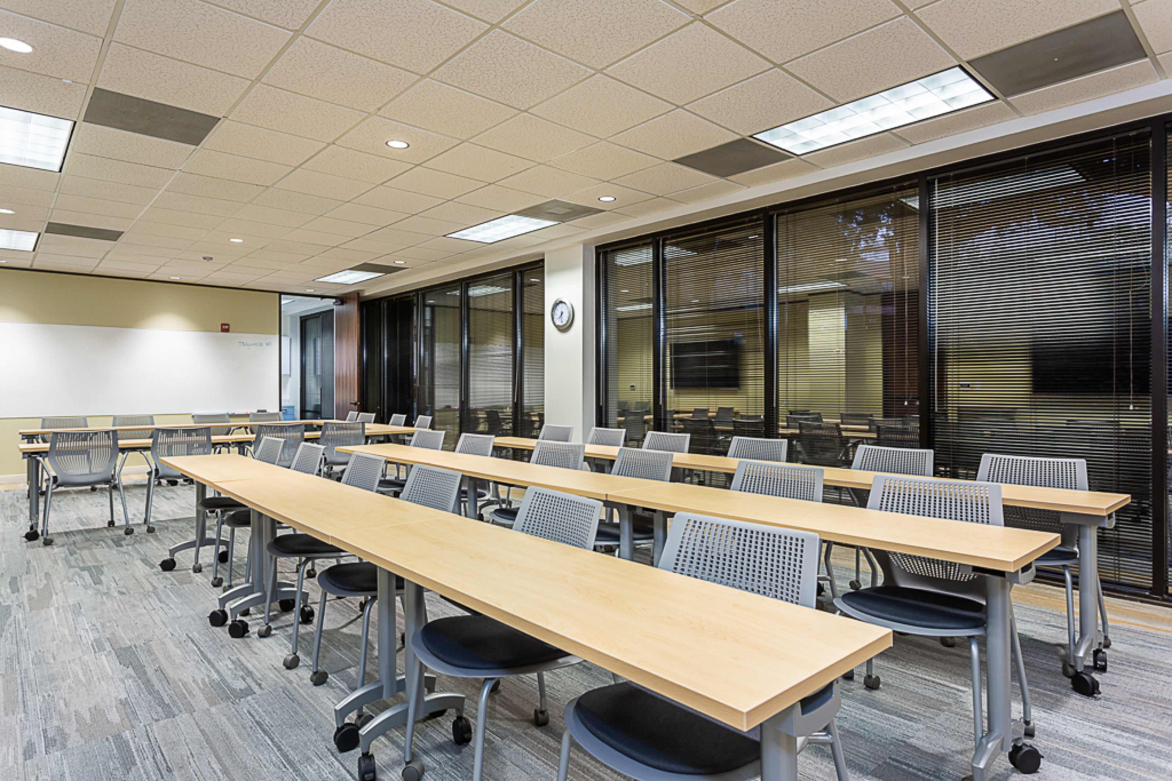 A spacious classroom features long tables arranged in a linear layout, equipped with ergonomic chairs and large windows illuminating the space.