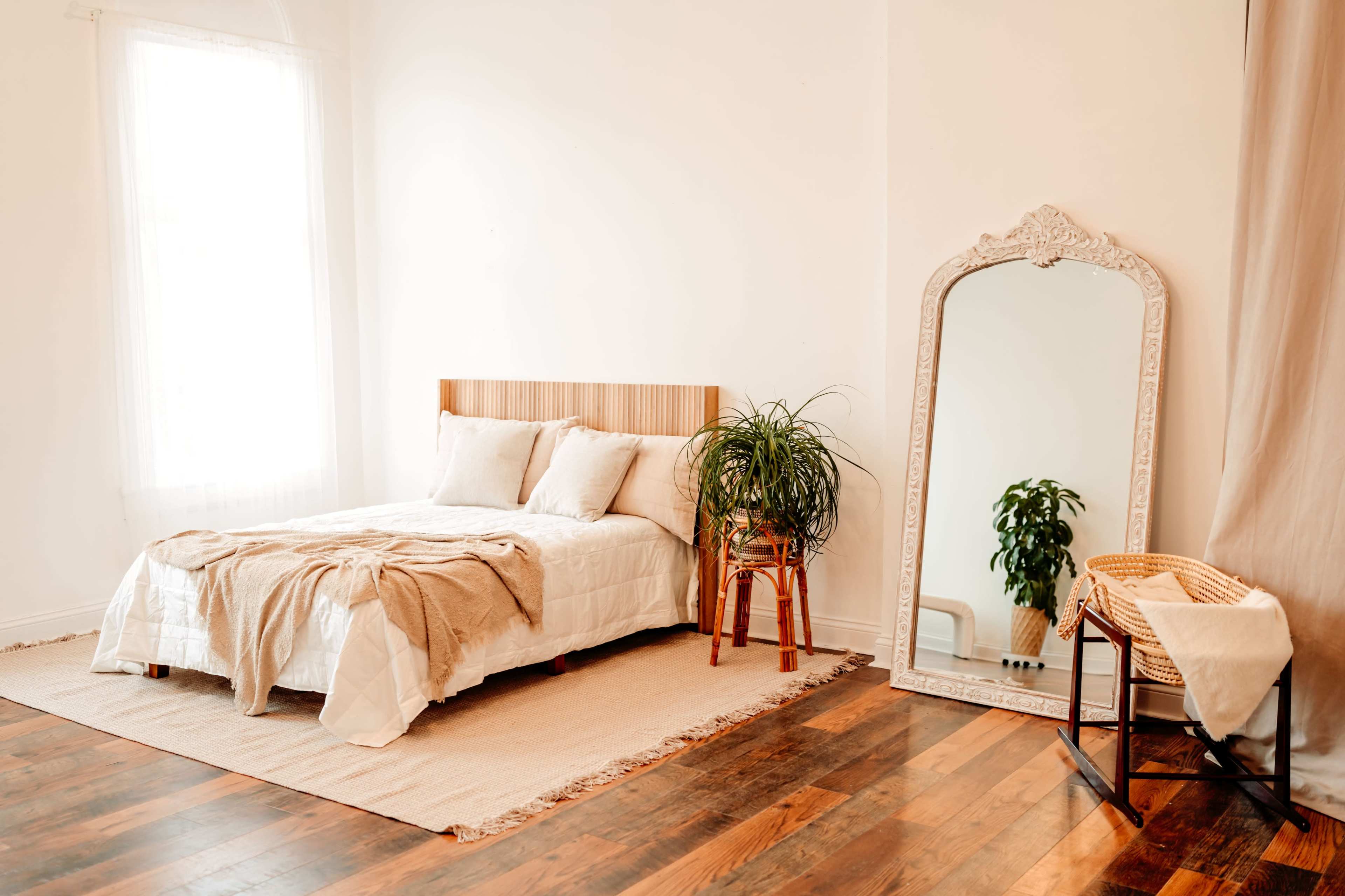 A cozy bedroom with a wooden bed, a large mirror, a plant on a stand, and a woven basket next to a light-colored rug on a hardwood floor.