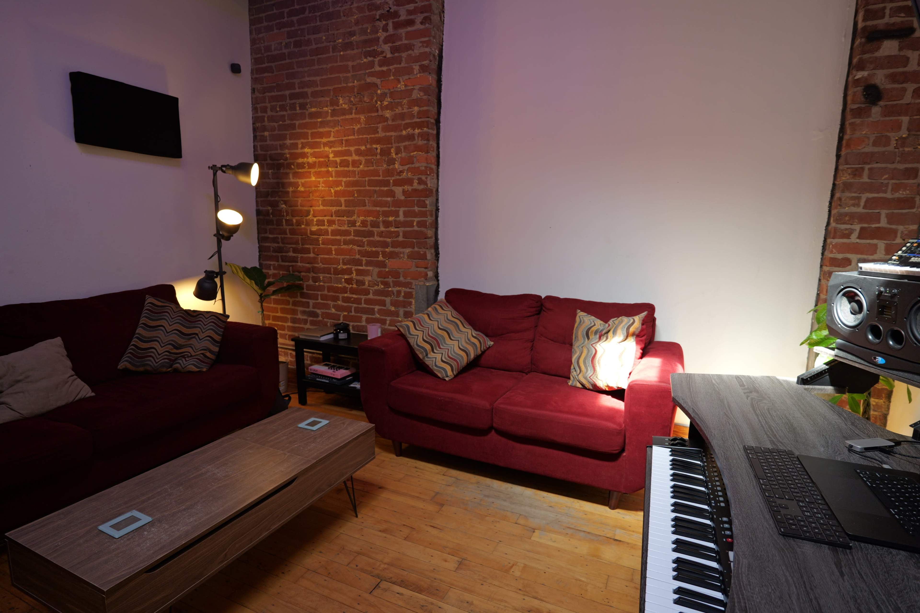 The scene features a cozy living room with red sofas, a wooden coffee table, and musical equipment on a desk against a backdrop of exposed brick walls.