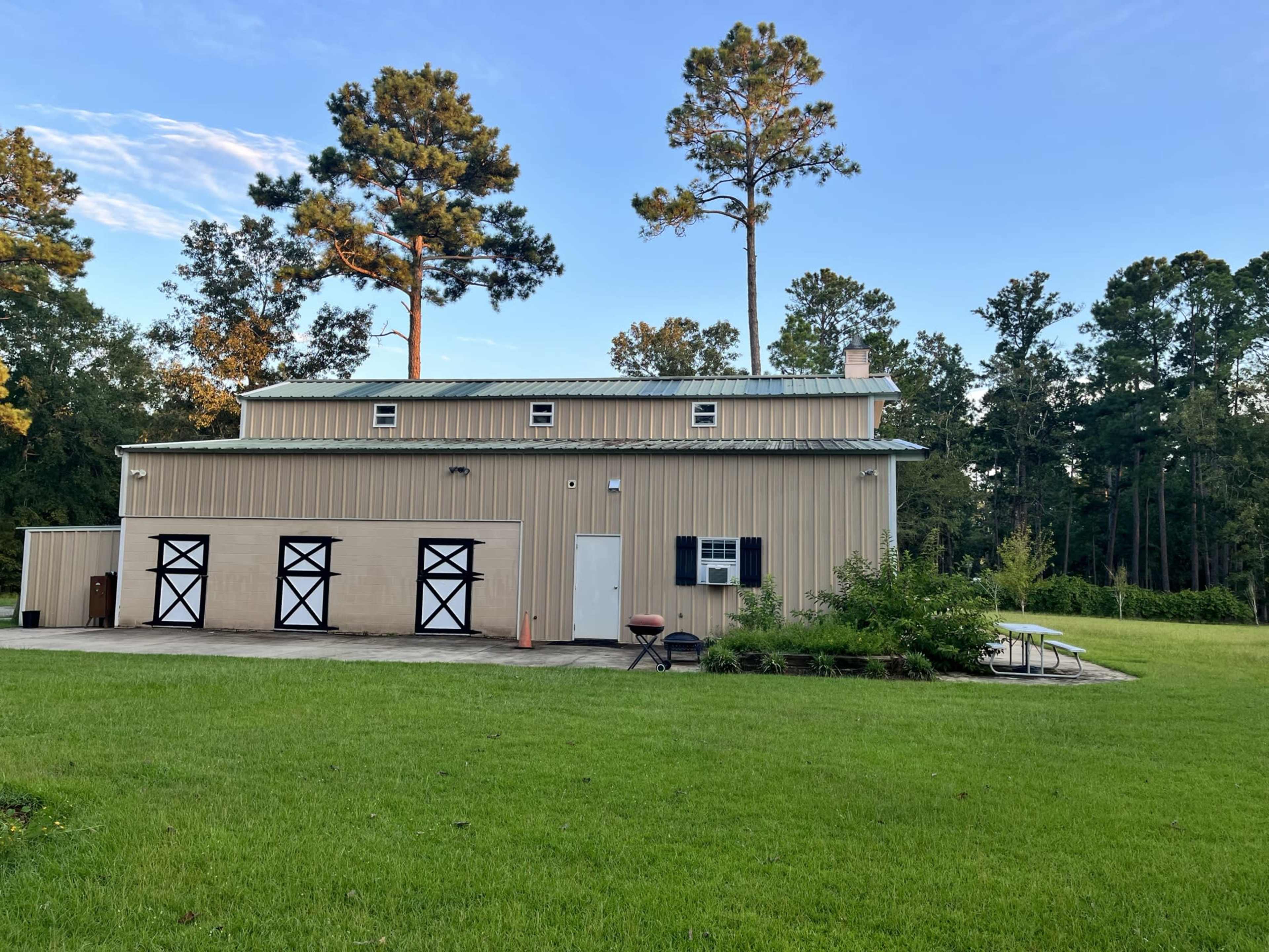 A large metal building with a tan exterior and three black garage doors is situated on a green lawn surrounded by trees.