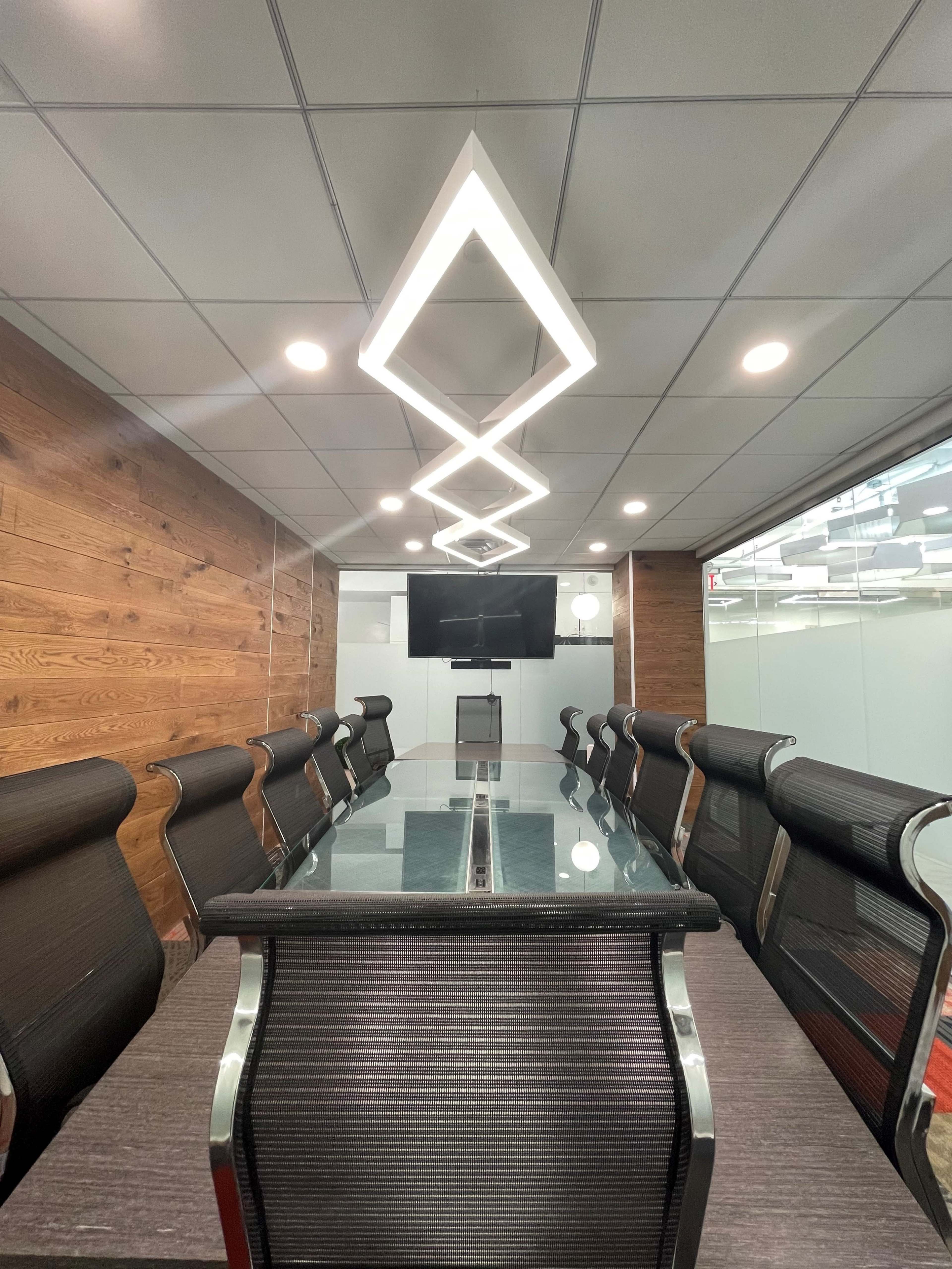 A modern conference room features a long glass table surrounded by black mesh chairs, with sleek pendant lighting and a TV mounted on the wall.