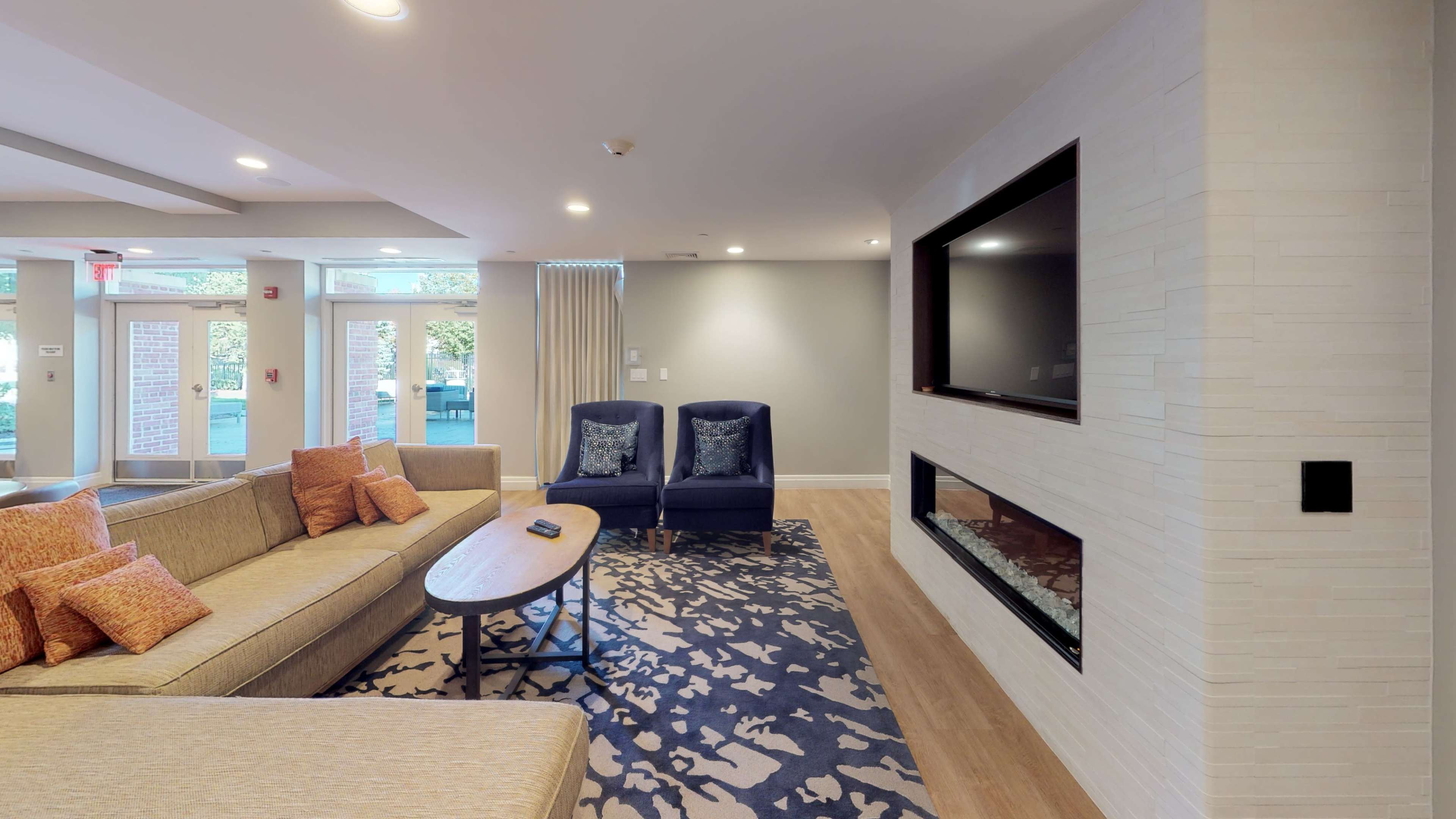 A modern living room with a beige sofa, a wooden coffee table, two blue chairs, and a wall-mounted television above a low fireplace.