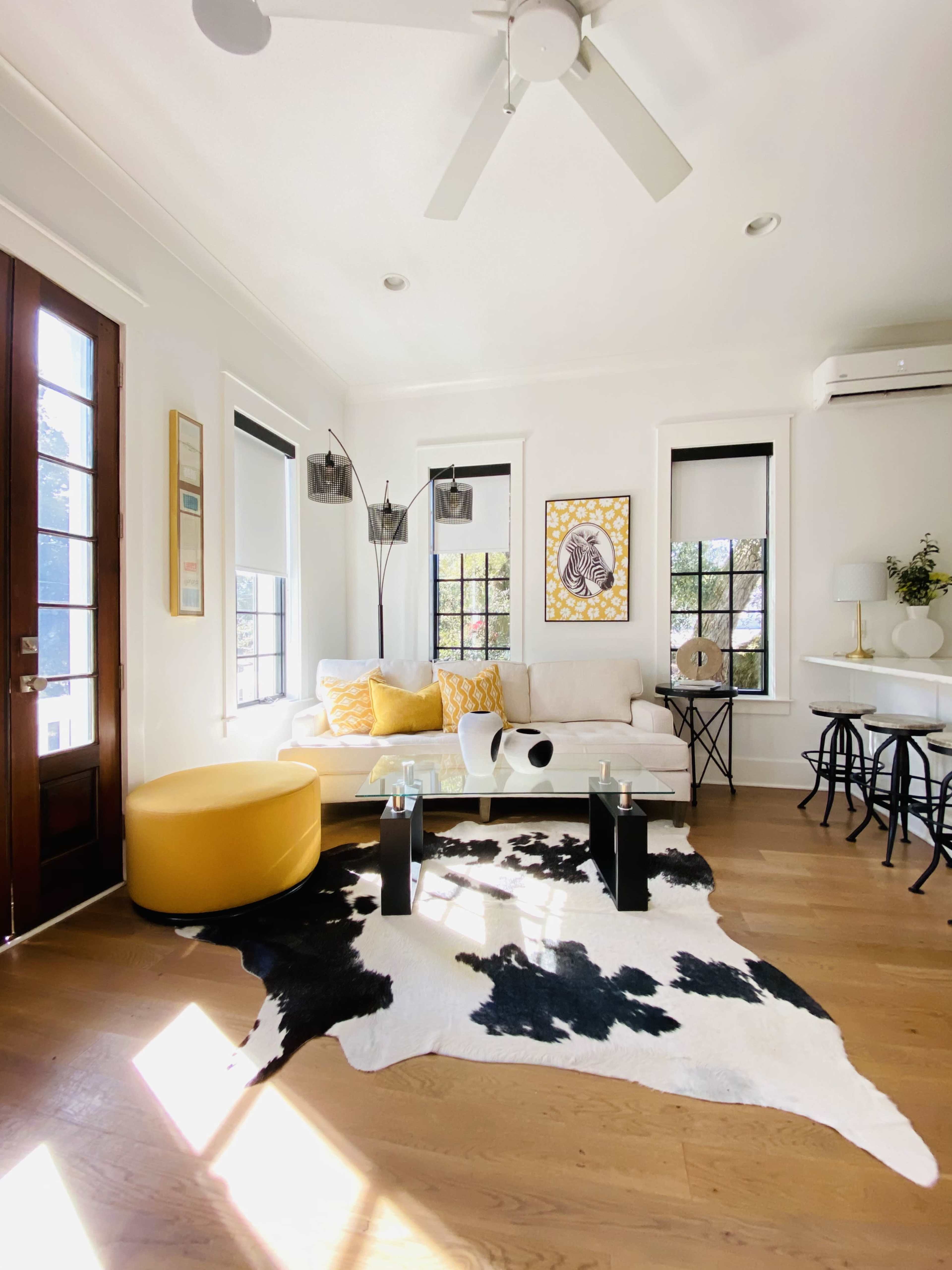 A bright living room features a white sofa with yellow accents, a cowhide rug on the wooden floor, and large windows allowing natural light.