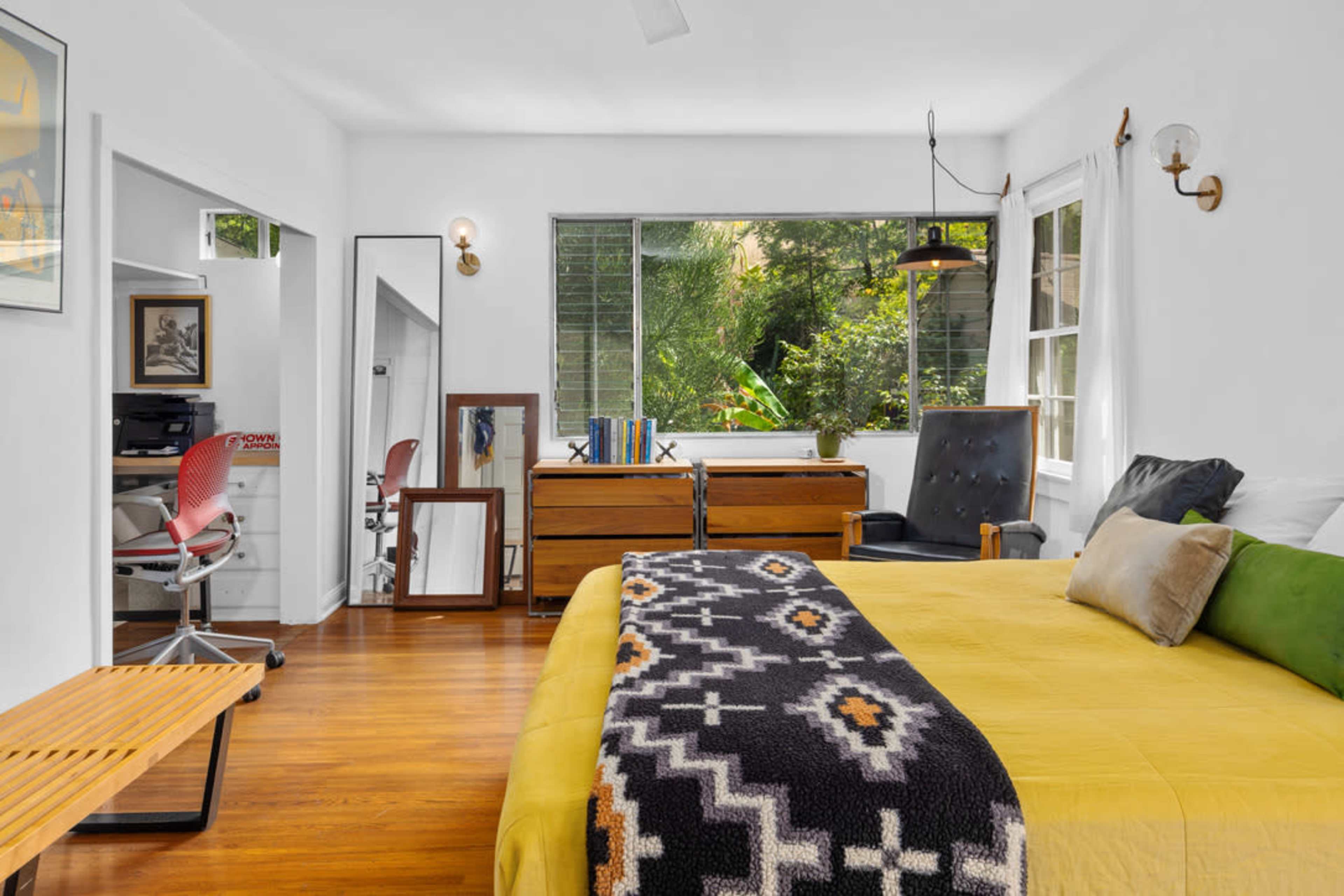 The image shows a bright bedroom featuring a yellow bedspread, a wooden dresser, a chair, and large windows allowing natural light to illuminate the space.