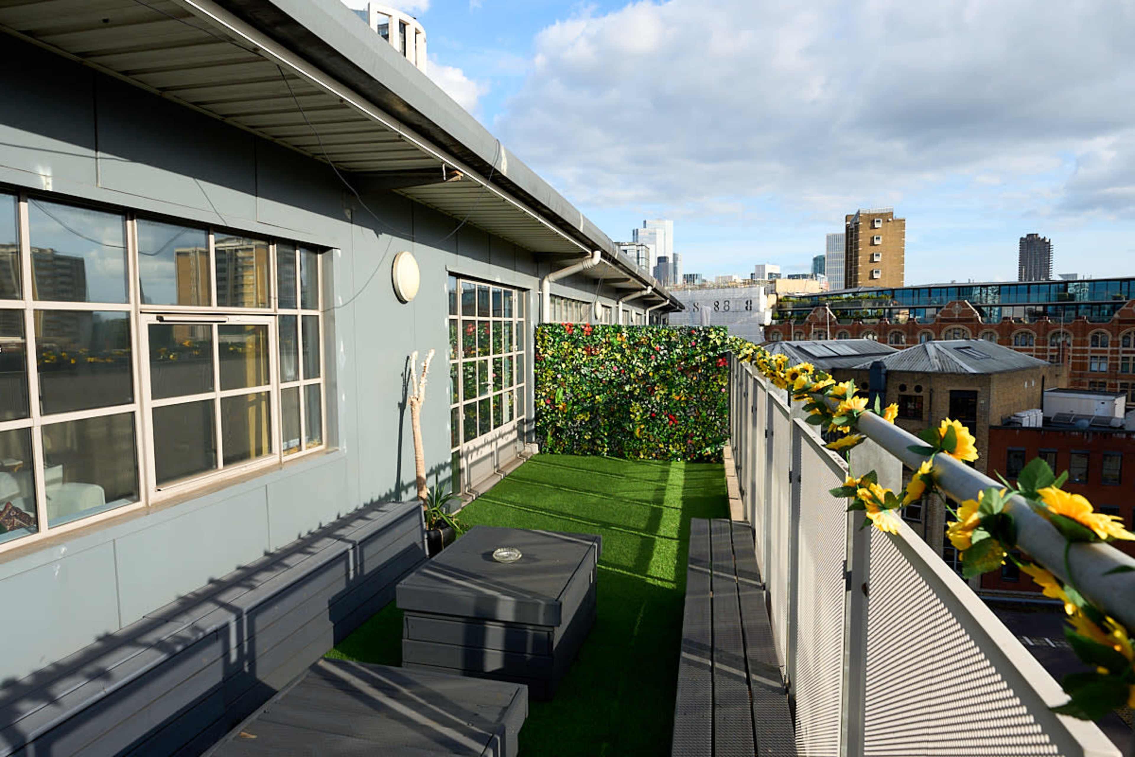 The image shows a rooftop terrace with green artificial grass, decorative foliage, and seating areas overlooking a cityscape.