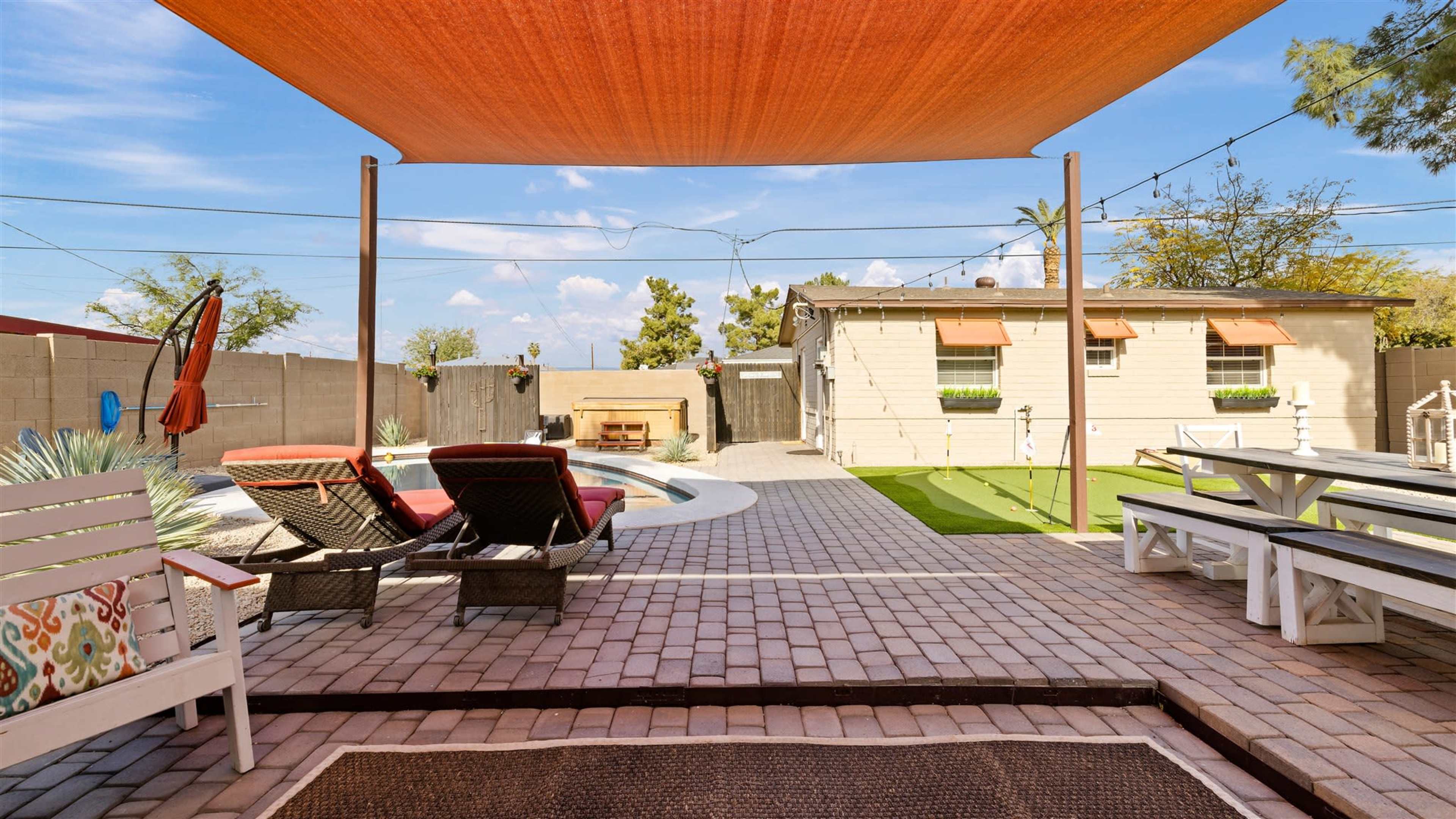 A backyard with a swimming pool, lounge chairs, a patio area, and a covered seating space, surrounded by a brick pathway and landscaped greenery.