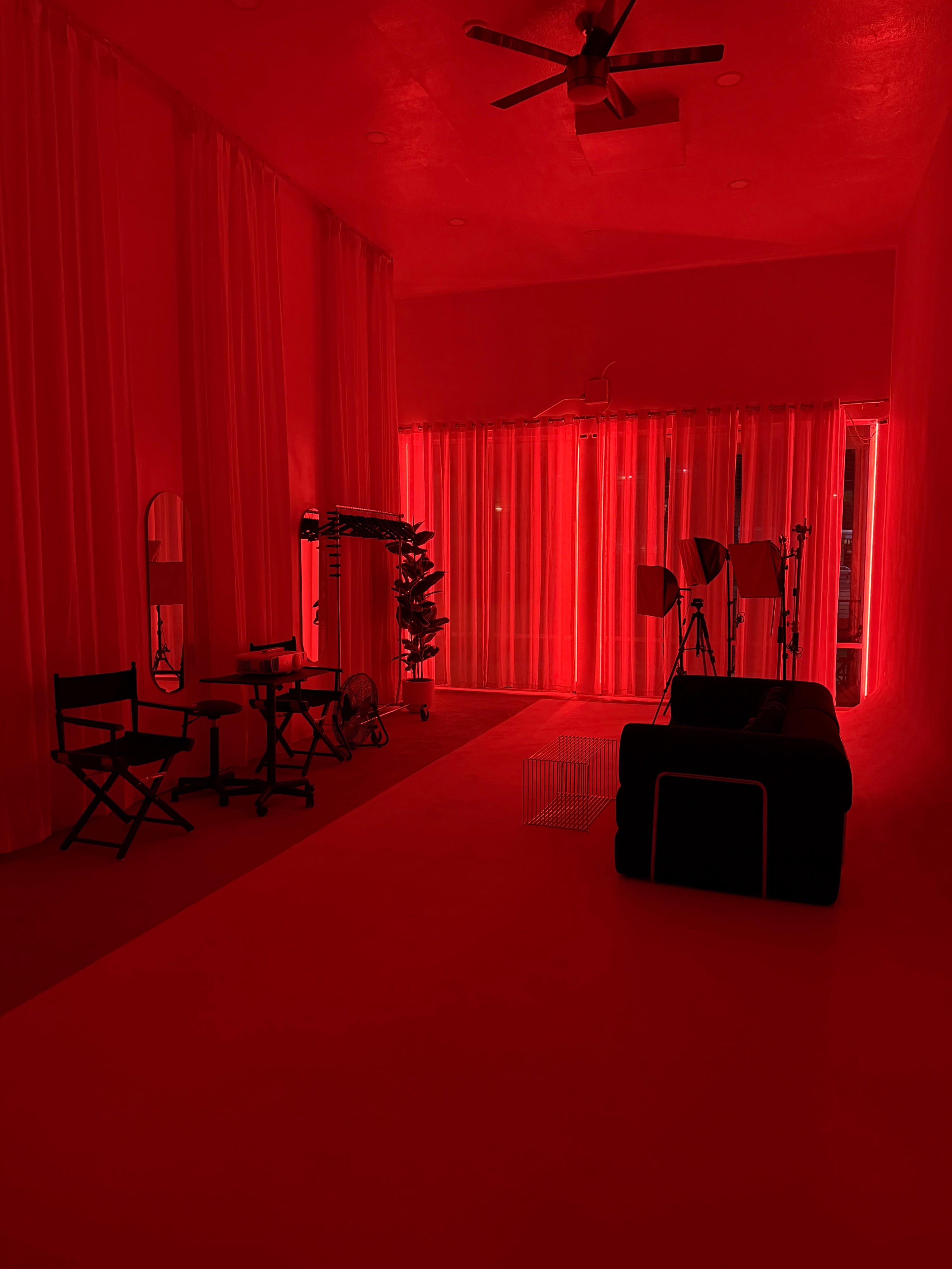 The image shows a minimalist room illuminated with red lighting, featuring a black couch, director's chairs, and equipment stands against white curtains.