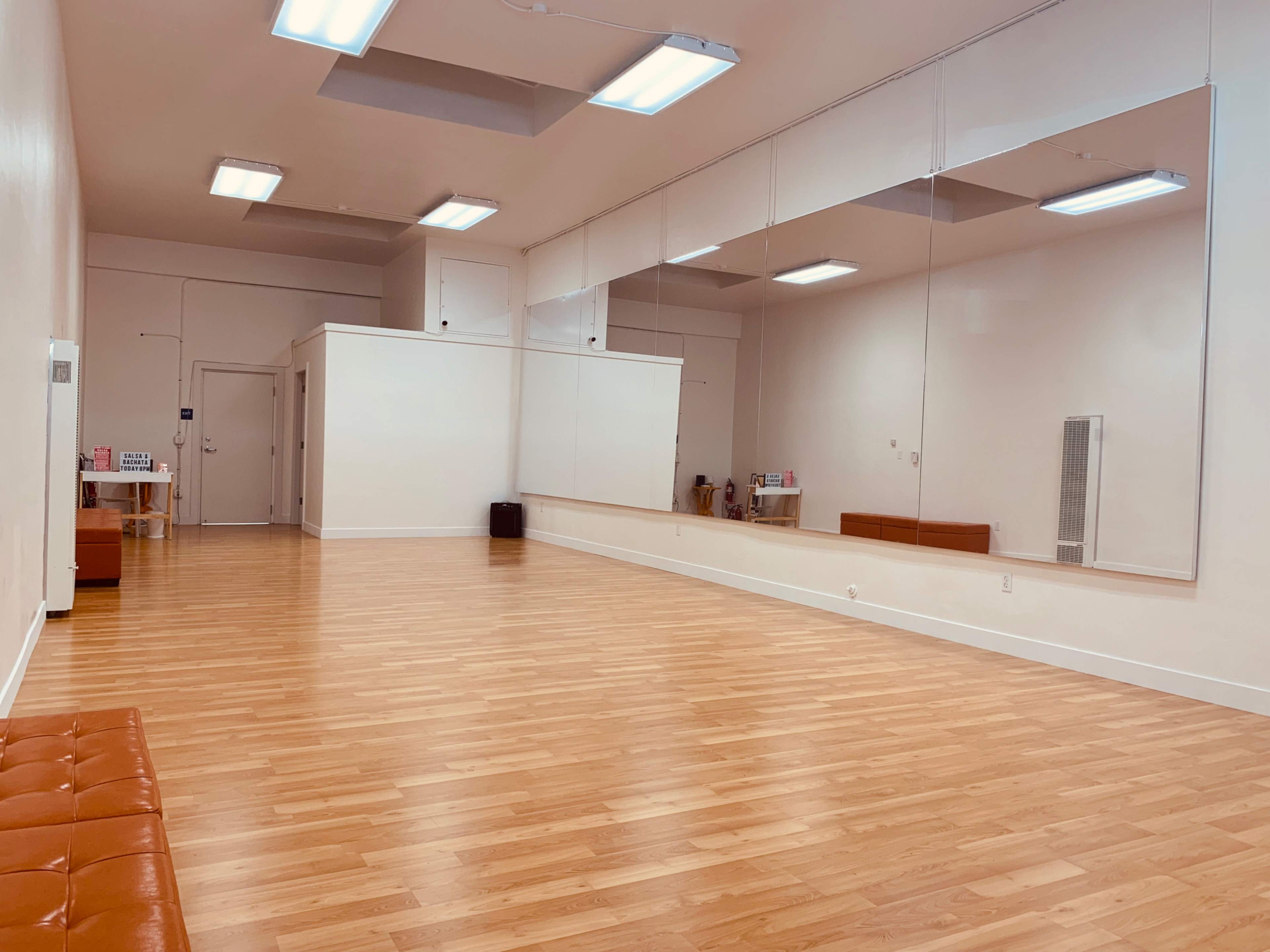 An empty dance studio with wooden flooring, large mirrors on one wall, and bright overhead lights.
