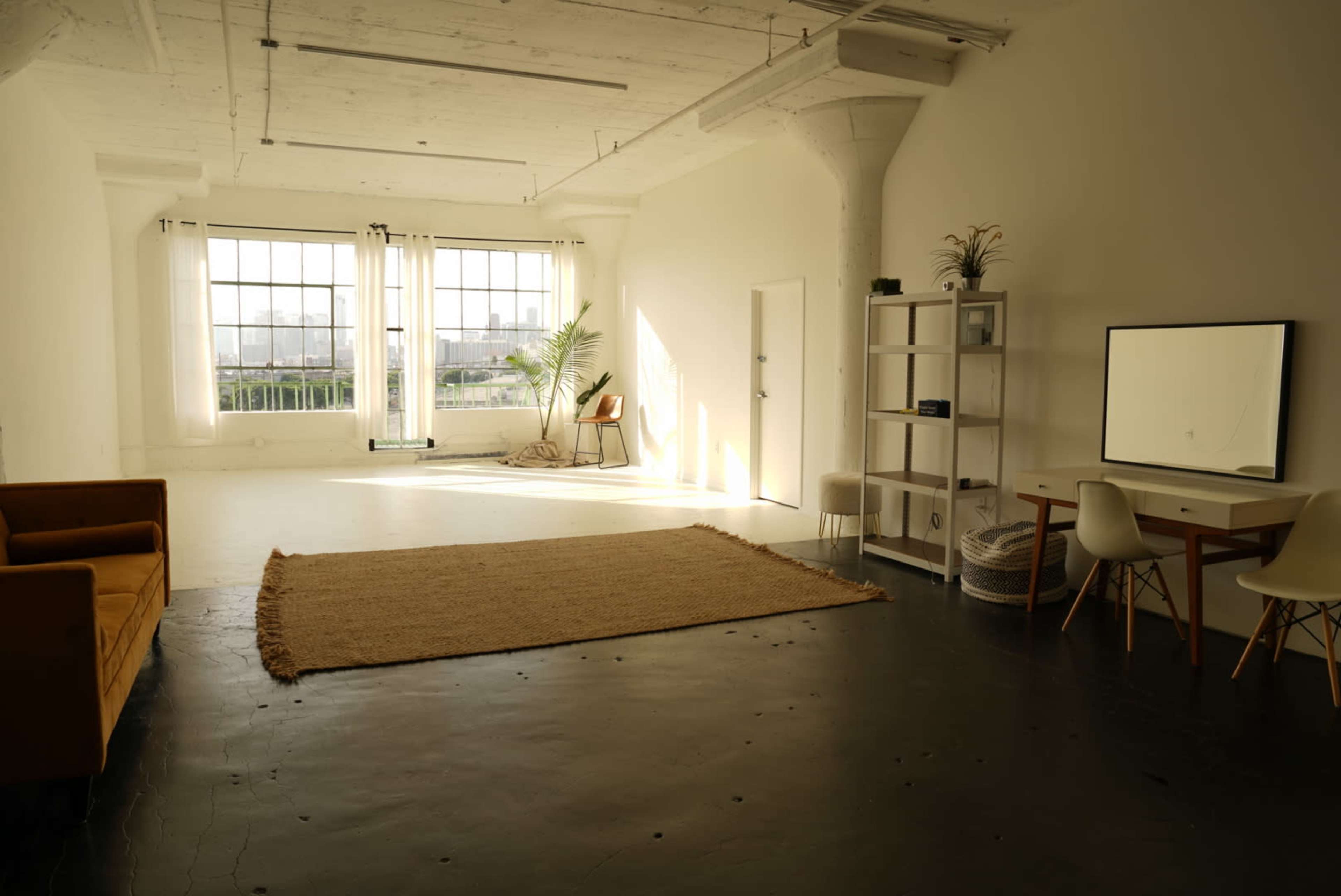 A spacious, well-lit room with large windows, a couch, a table with a mirror, and minimal decor.