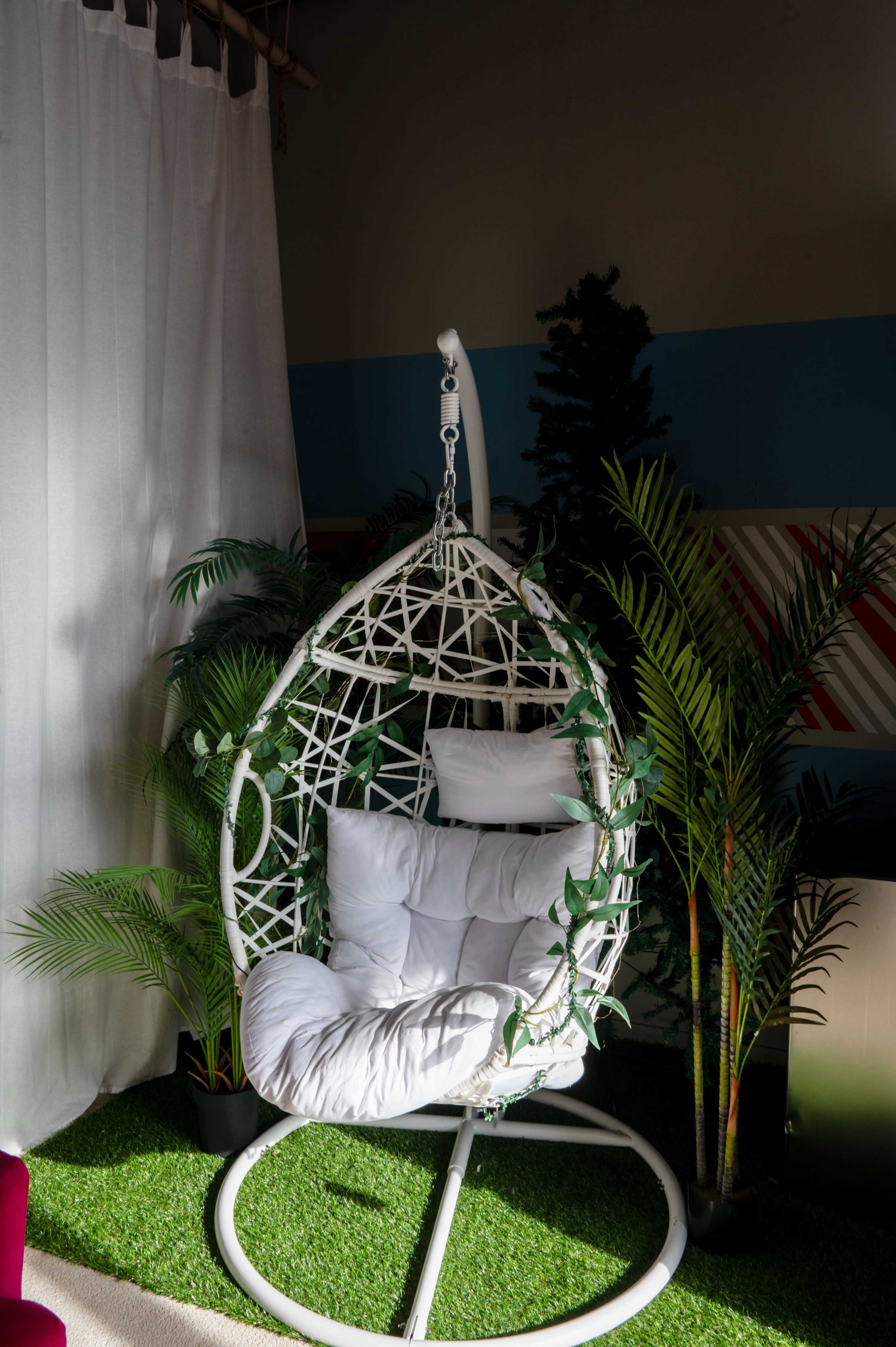 A white hanging chair with cushions is surrounded by indoor plants and artificial grass in a brightly lit room.
