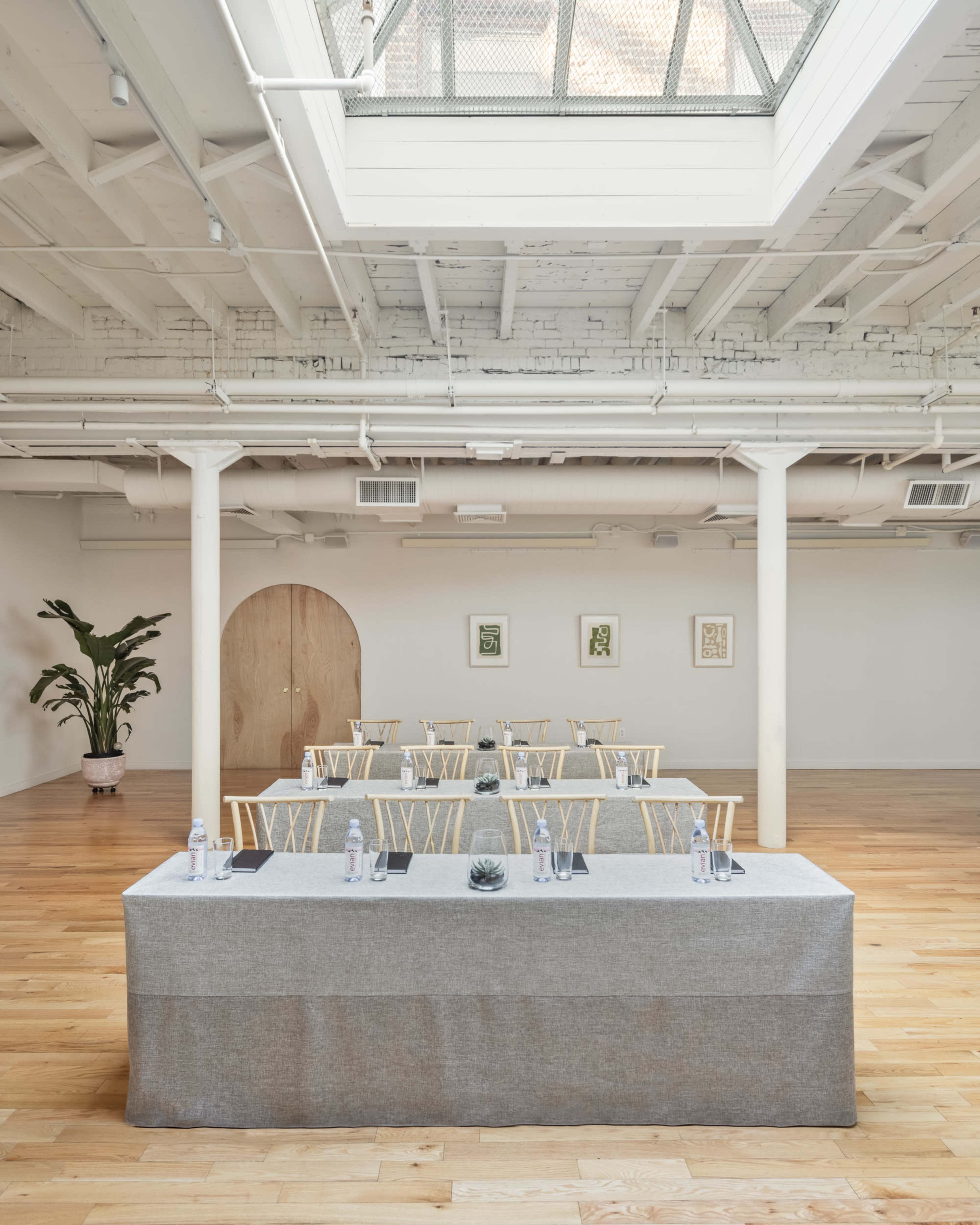 A conference room features a central table with water bottles and notepads, surrounded by empty chairs and adorned with a plant and artwork.