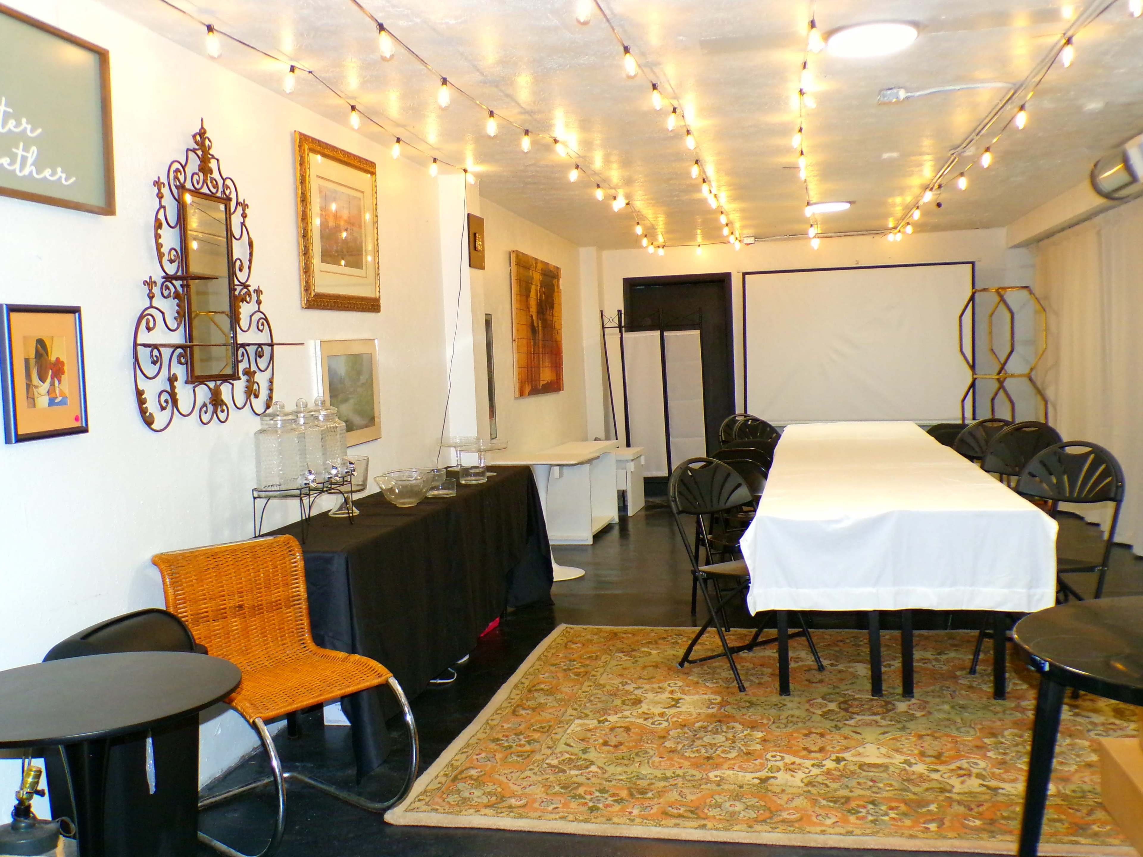 The image shows a decorated room with a long table covered in a white tablecloth, surrounded by black chairs, and illuminated by string lights overhead.