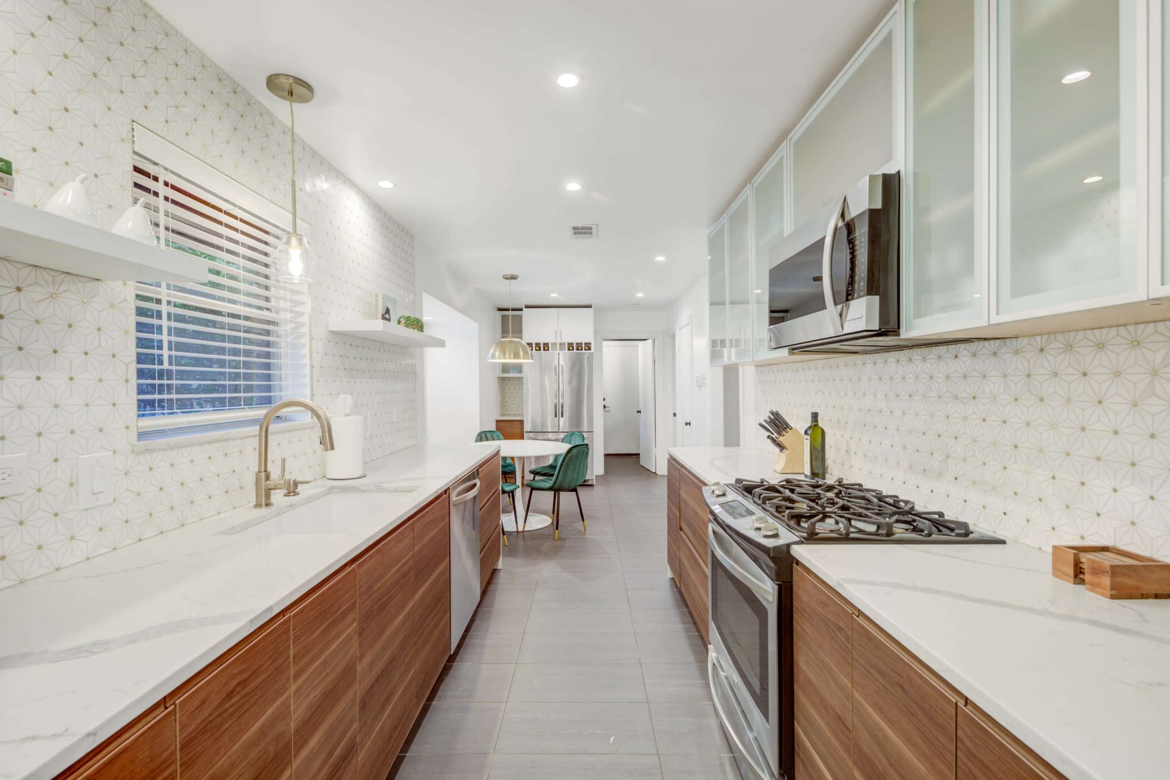 A modern kitchen features wooden cabinetry, marble countertops, and stainless steel appliances, with a dining area visible through a doorway.