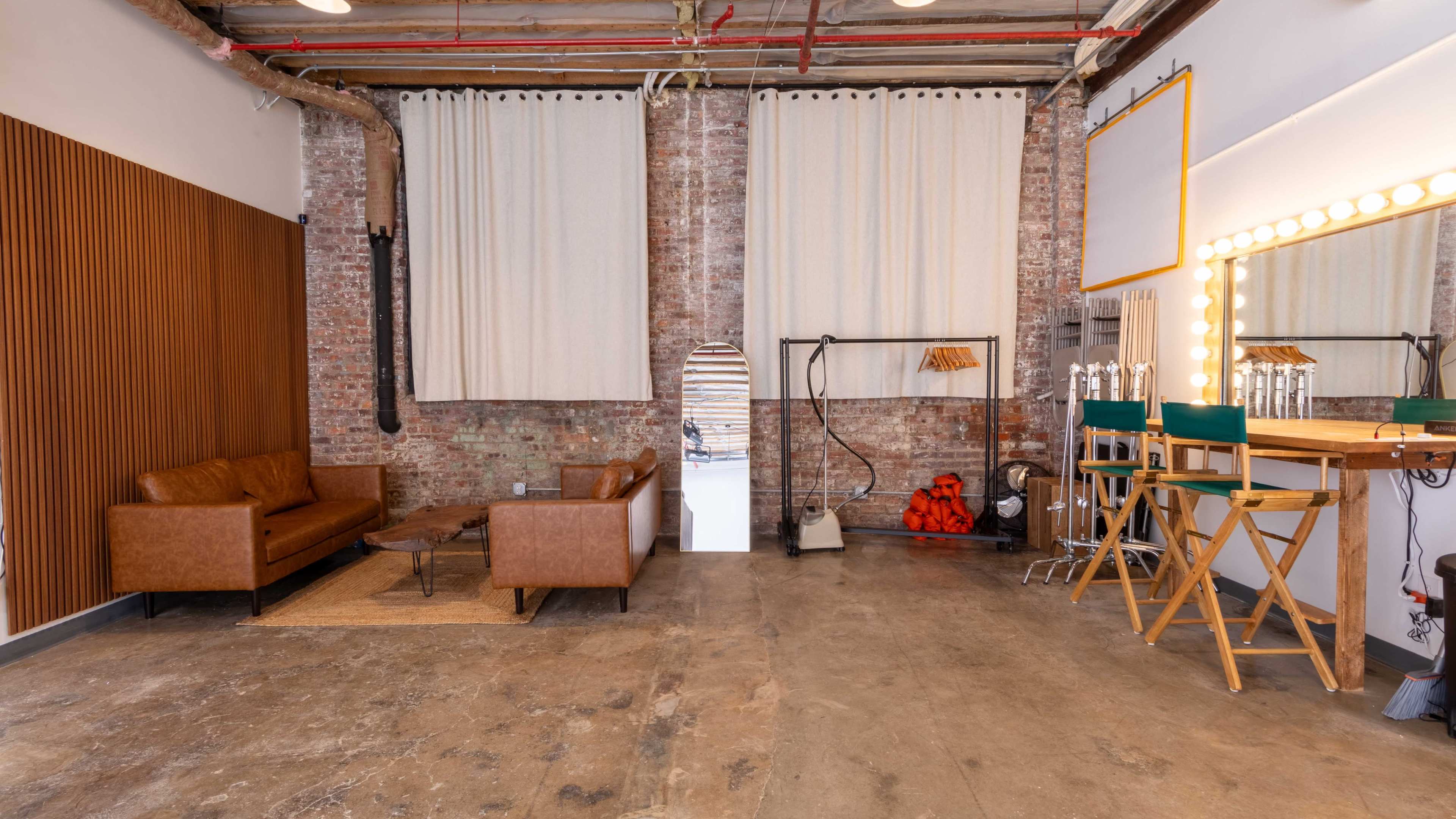 The image shows an industrial-style studio space featuring a brown leather couch, a wooden coffee table, a dressing area with lights, and an open doorway leading to another room.