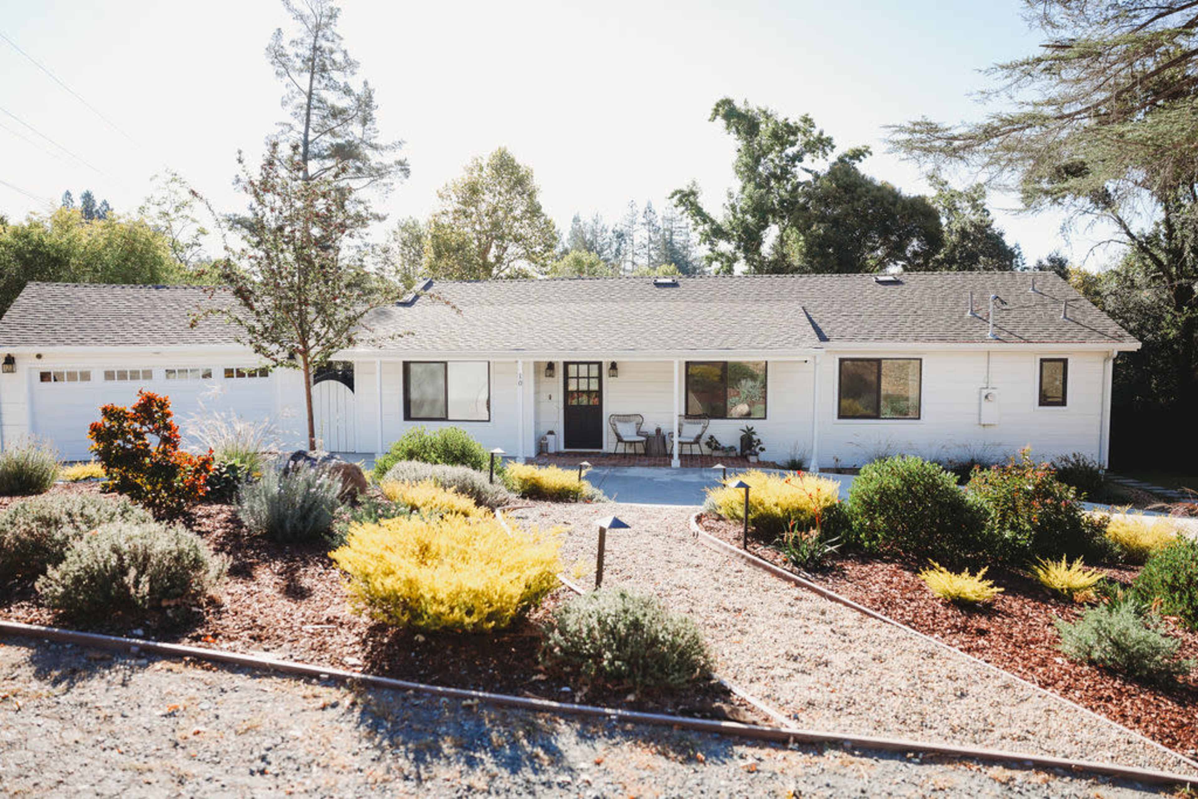 A single-story white house with a landscaped yard featuring a variety of shrubs and ornamental plants.