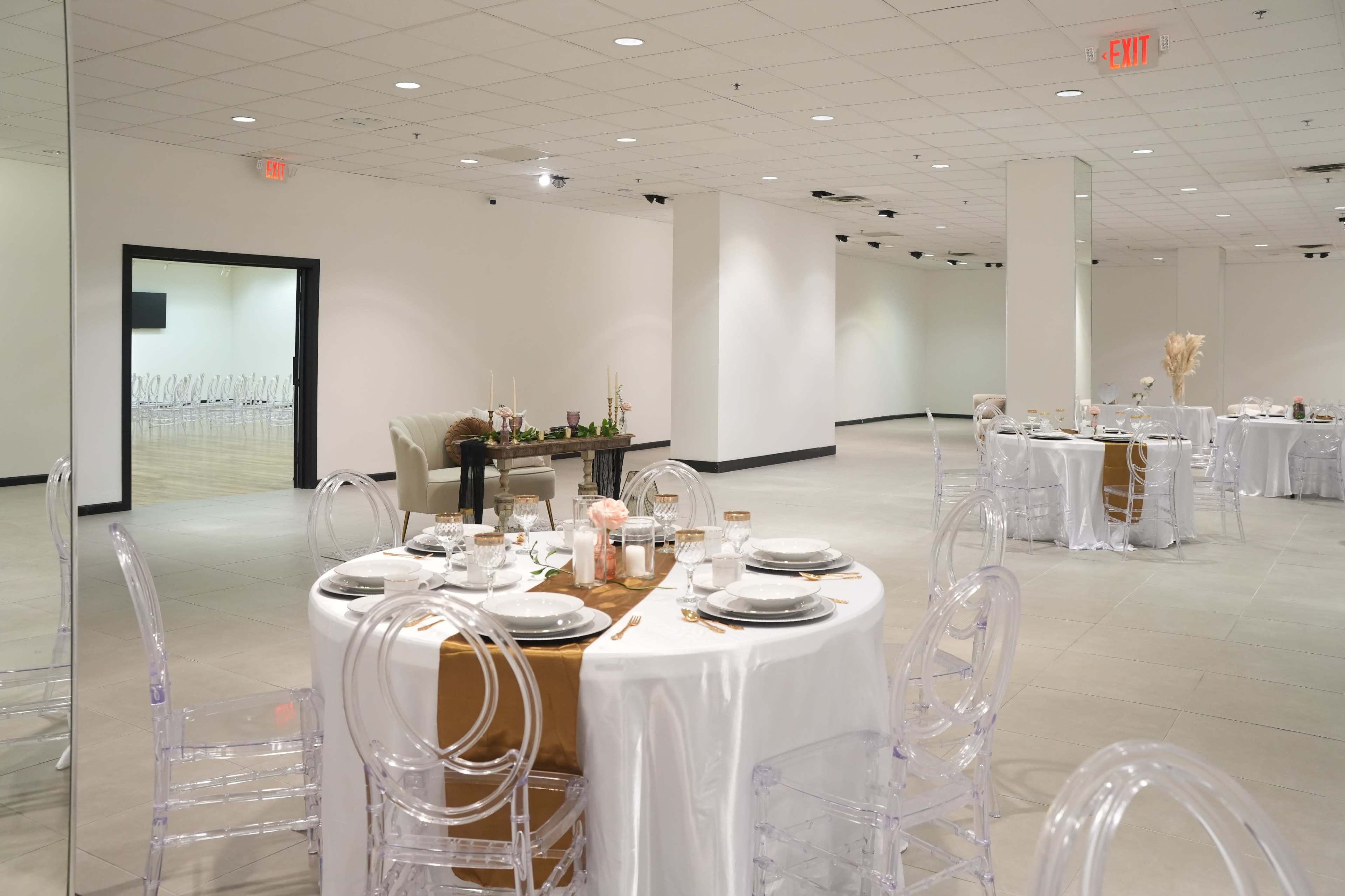A spacious event room features round tables set with white tablecloths and clear chairs, with a polished dance floor in the background.